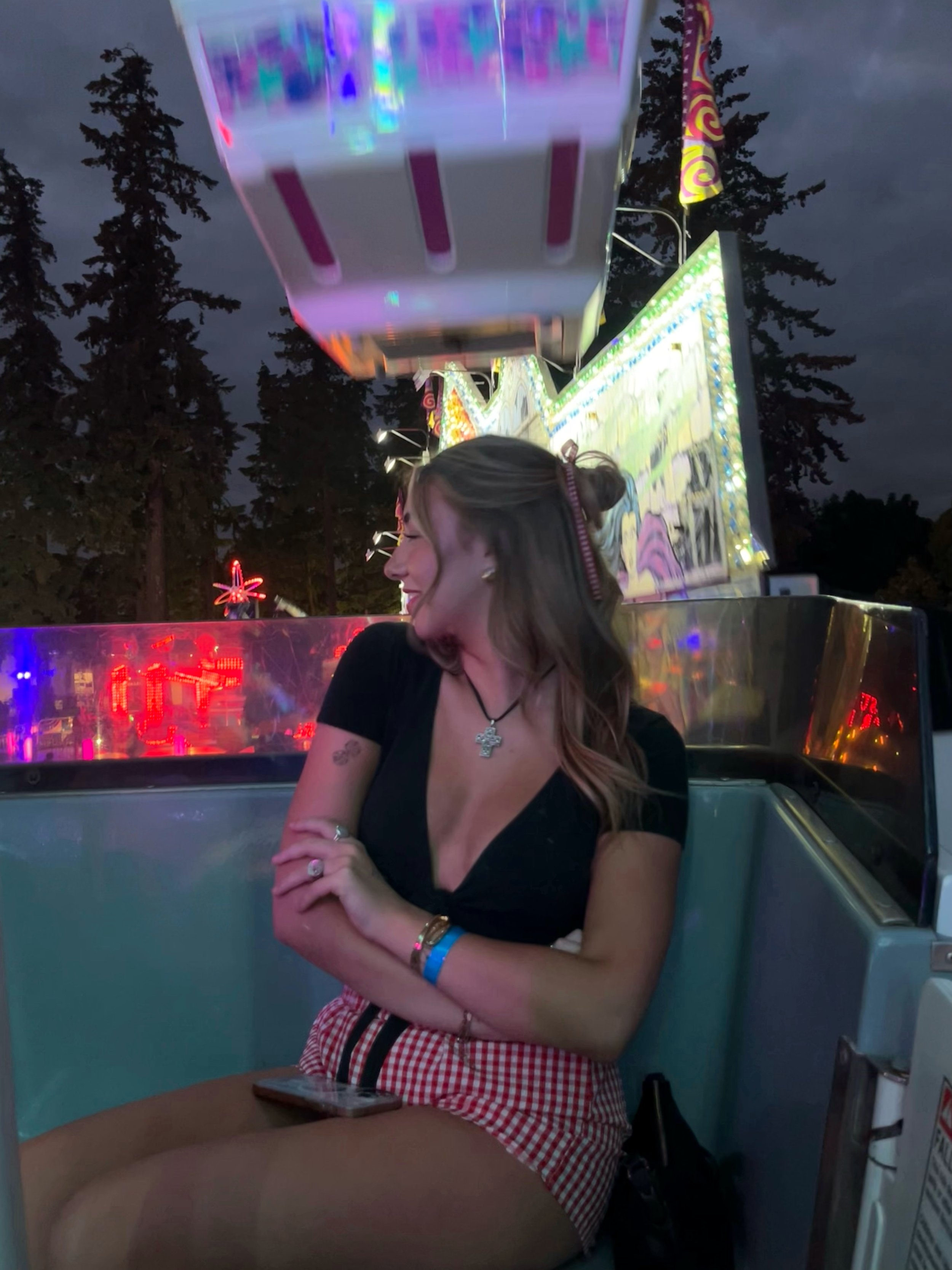 A young woman with light brown hair, wearing a black top, red checkered shorts, and jewelry, sitting in a carnival ride in the evening with amusement rides and bright lights in the background.