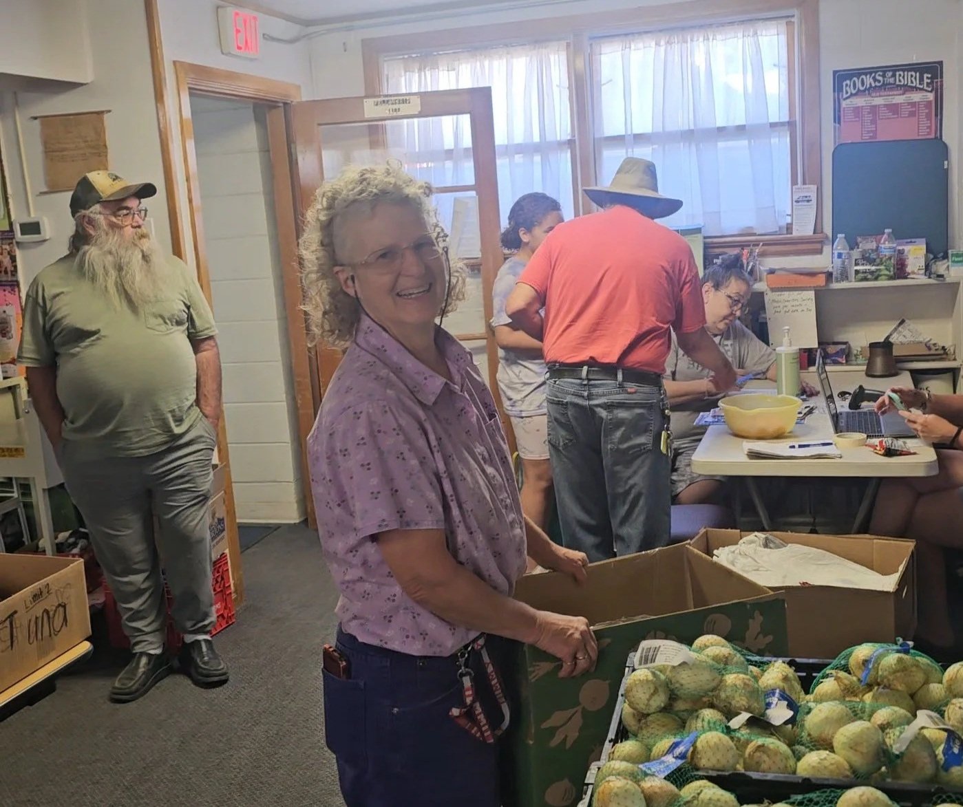 Group of people inside a room, with a woman in the foreground smiling at the camera, surrounded by boxes of produce, and others engaged in sorting or working at a table.