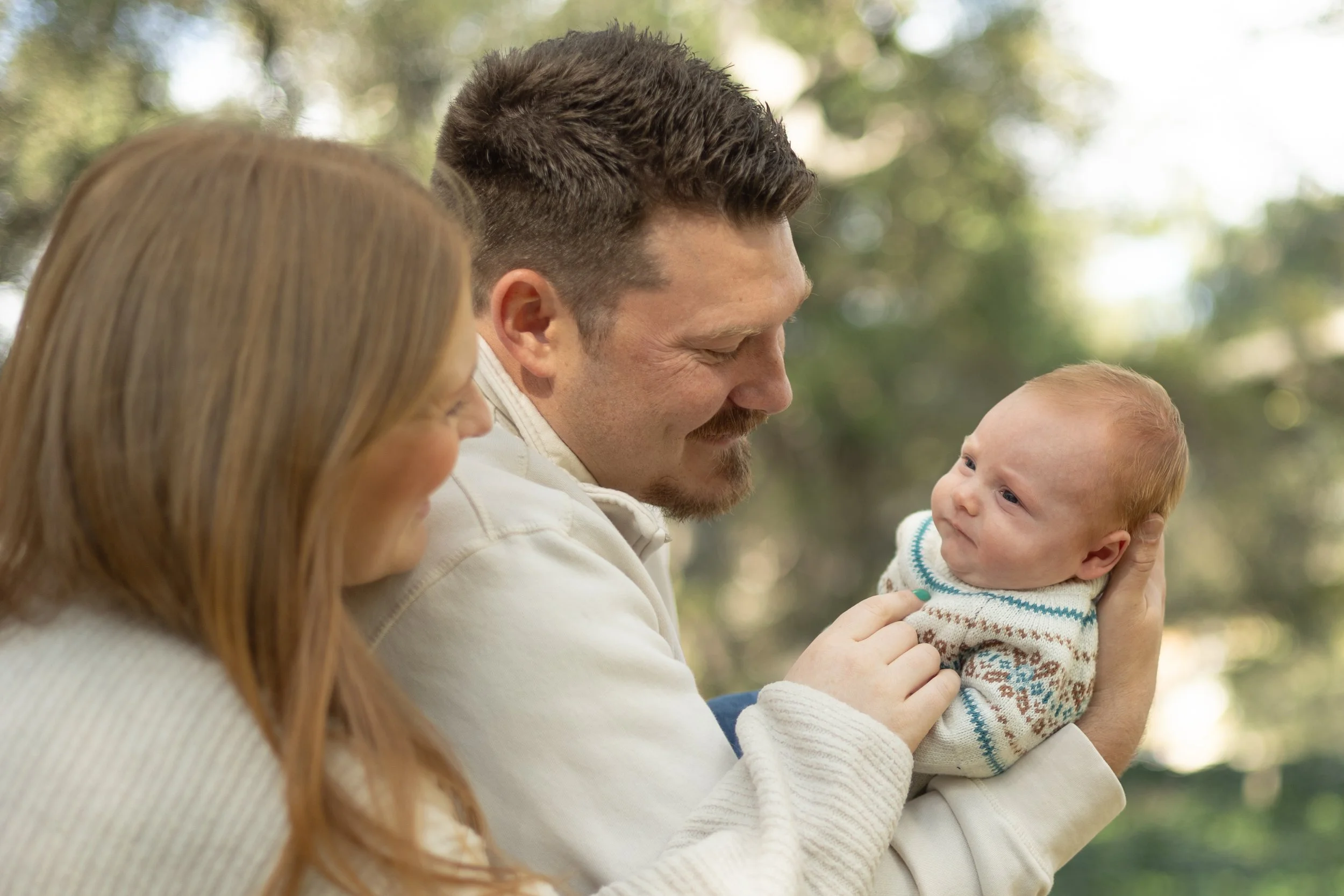 couple looking lovingly at their newborn while hugging in a park with trees in the background