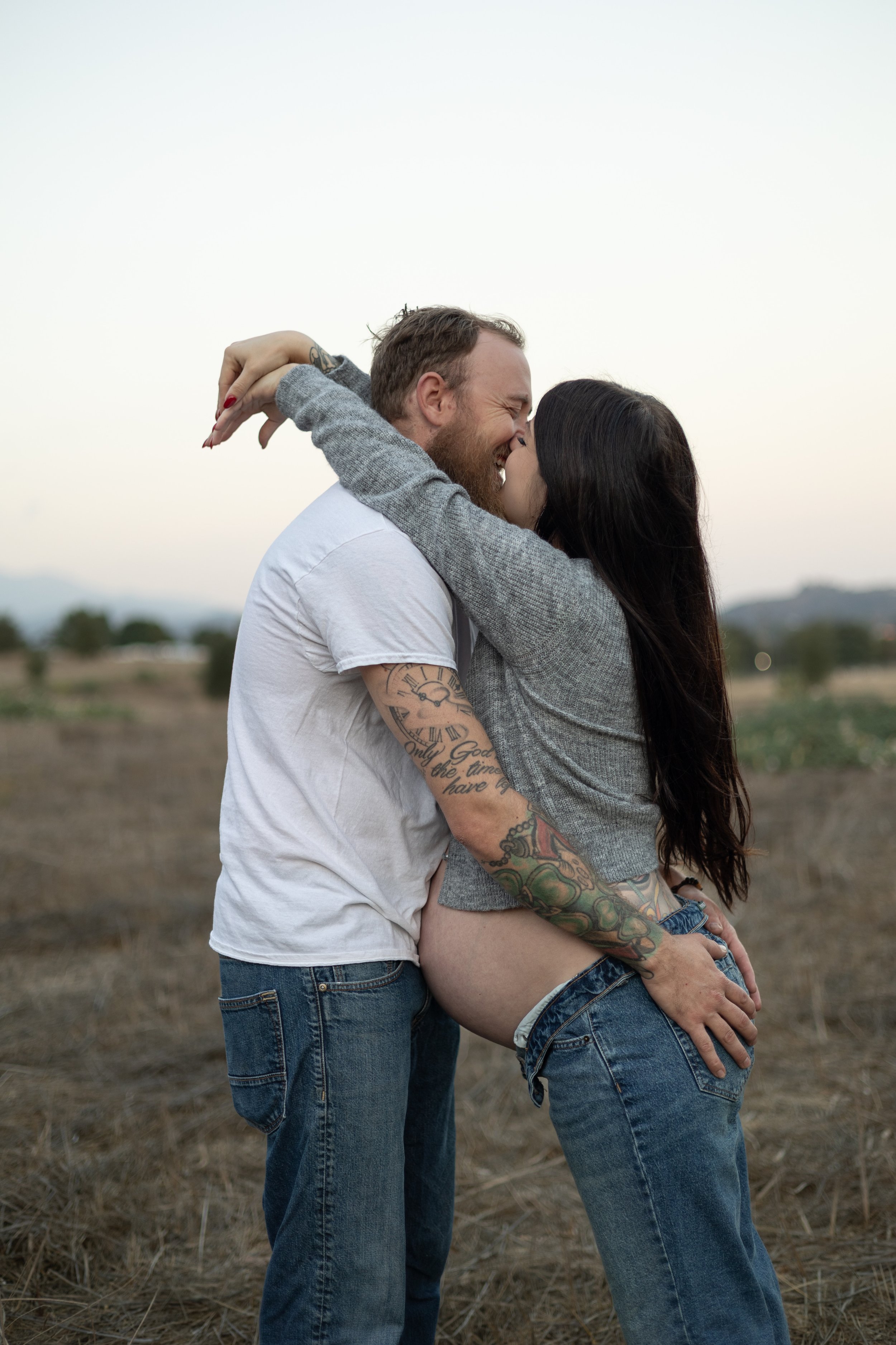 candid couple laughing and kissing in field with mountains in the background husband holding wife's pregnant belly