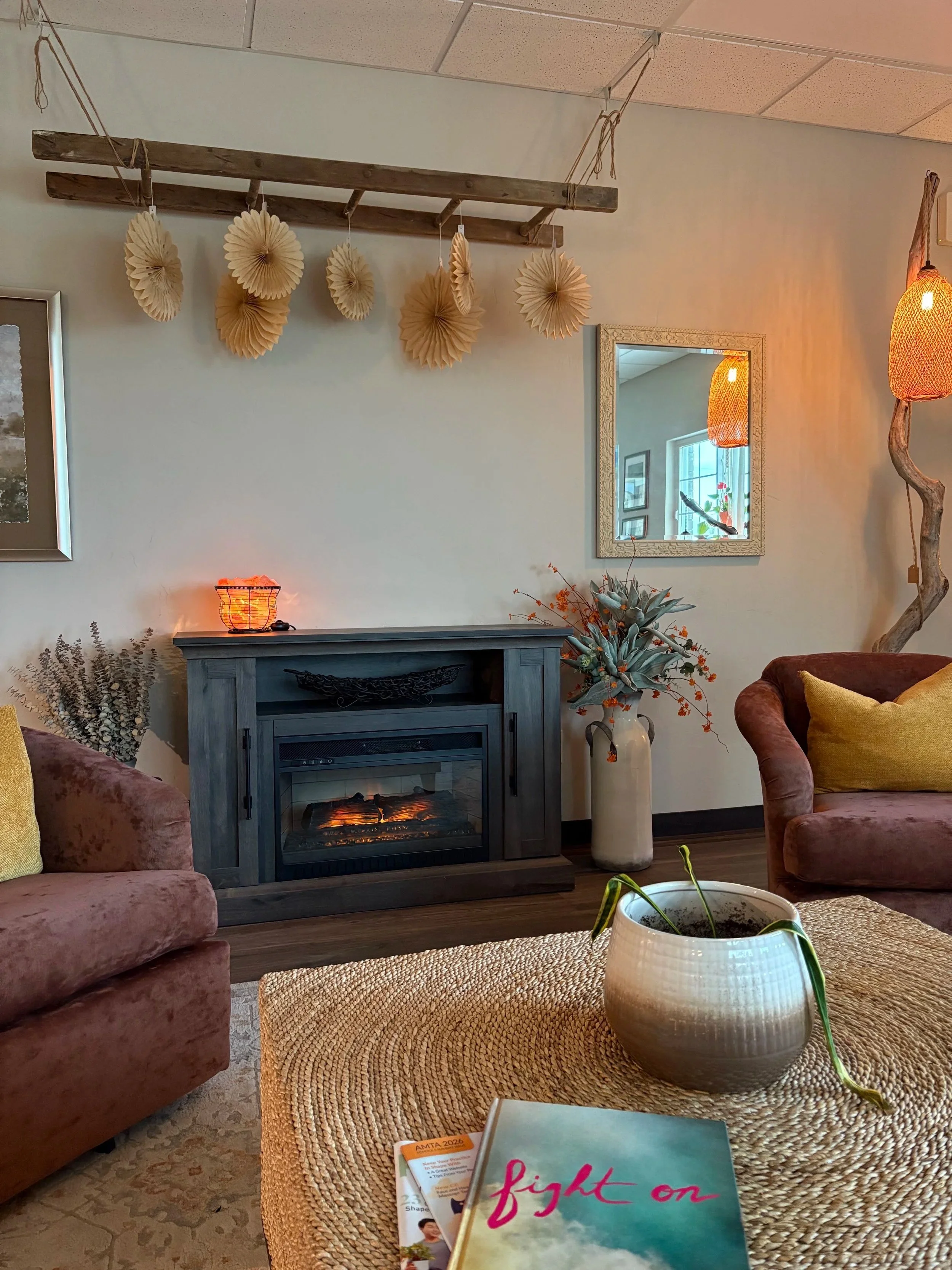 Living room with pink velvet chairs, a beige woven ottoman, a gray fireplace, a large beige vase with orange flowers, framed wall art, and a mirror reflecting a window with plants. Paper decorations hang from the ceiling, and a warm light emits from a lamp on a side table.