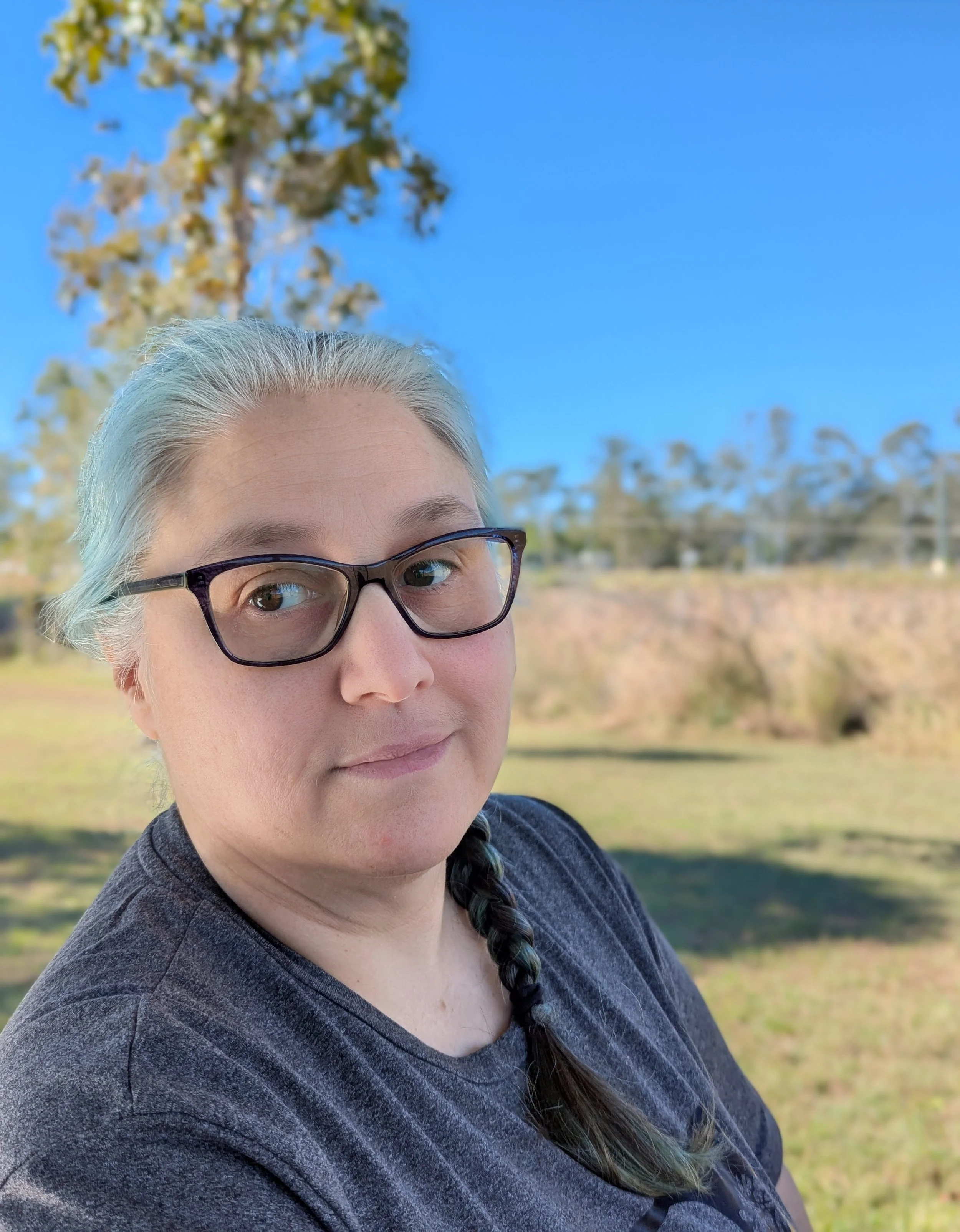 A woman with glasses and long hair in a braid standing outdoors on a sunny day, with trees and clear blue sky in the background.