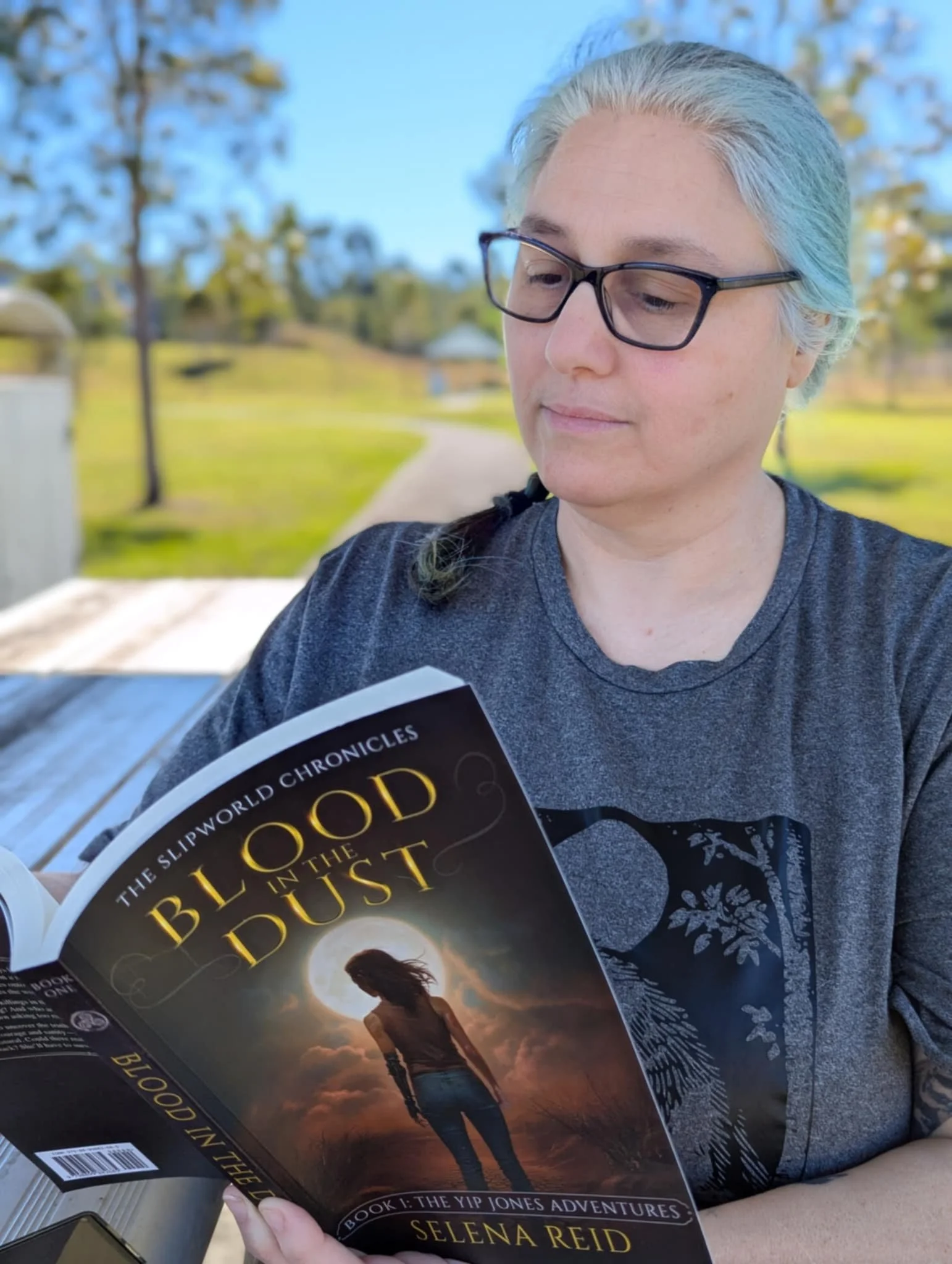 Person with blue-green hair and glasses reading a book titled 'Blood in the Dust' outdoors on a bench in a park.