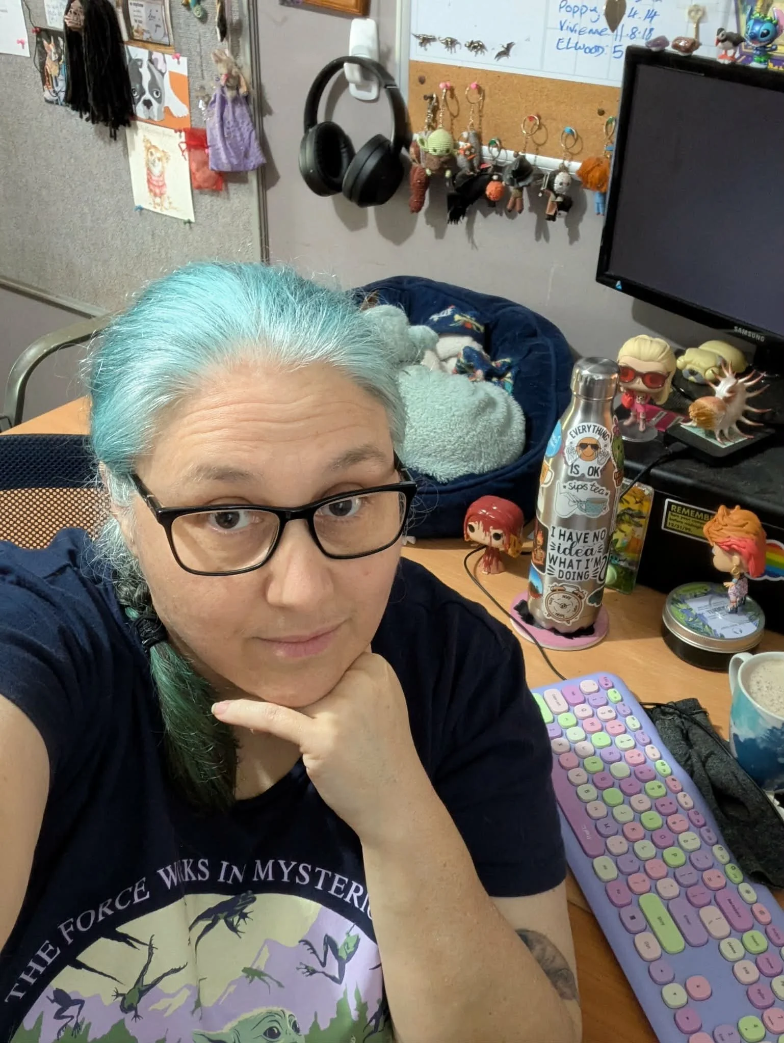 Person with glasses and blue-green hair taking a selfie at a cluttered desk in a home office or craft room.