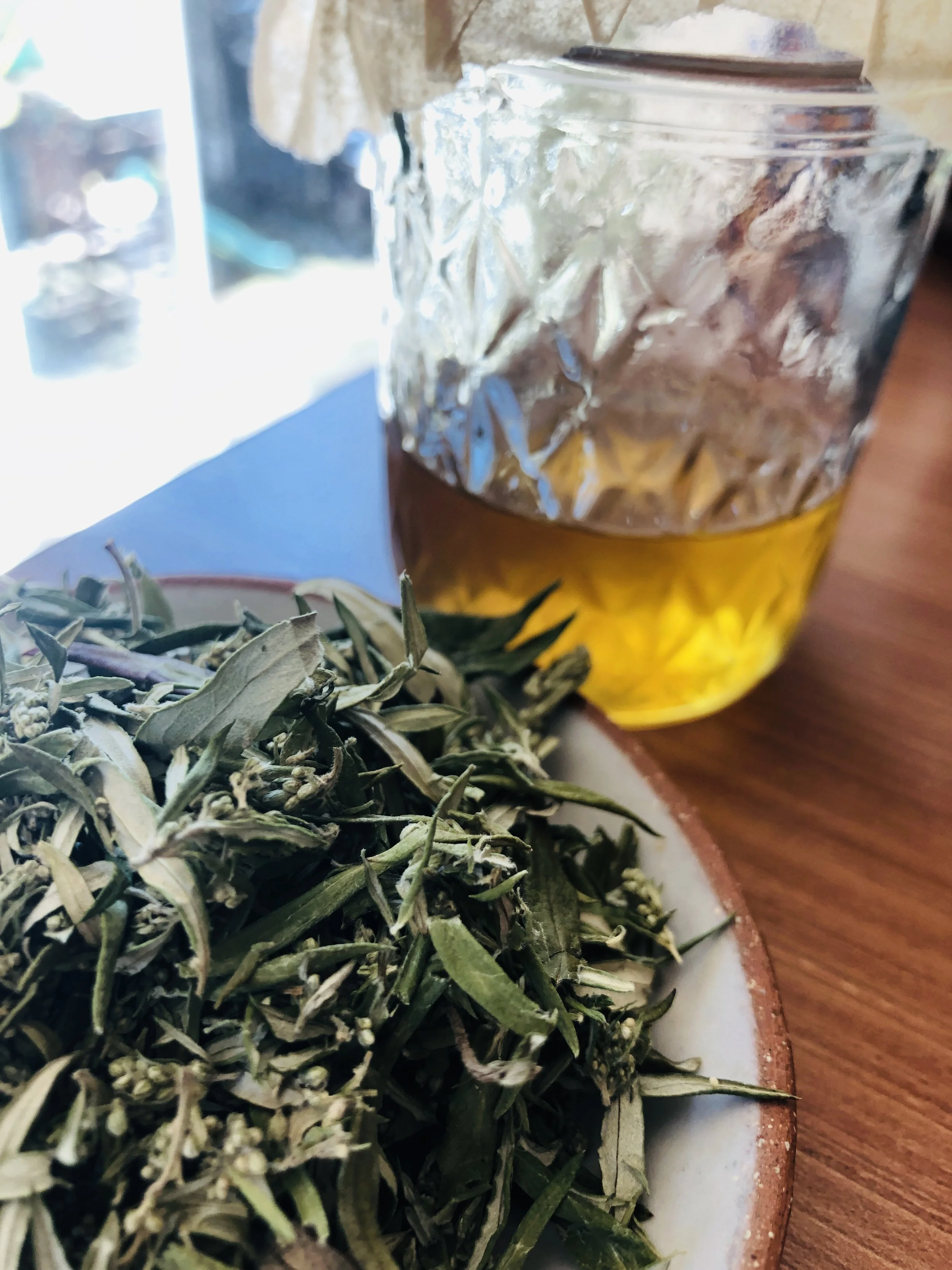 Close-up of dried green herbs on a white plate with a glass of tea in the background.