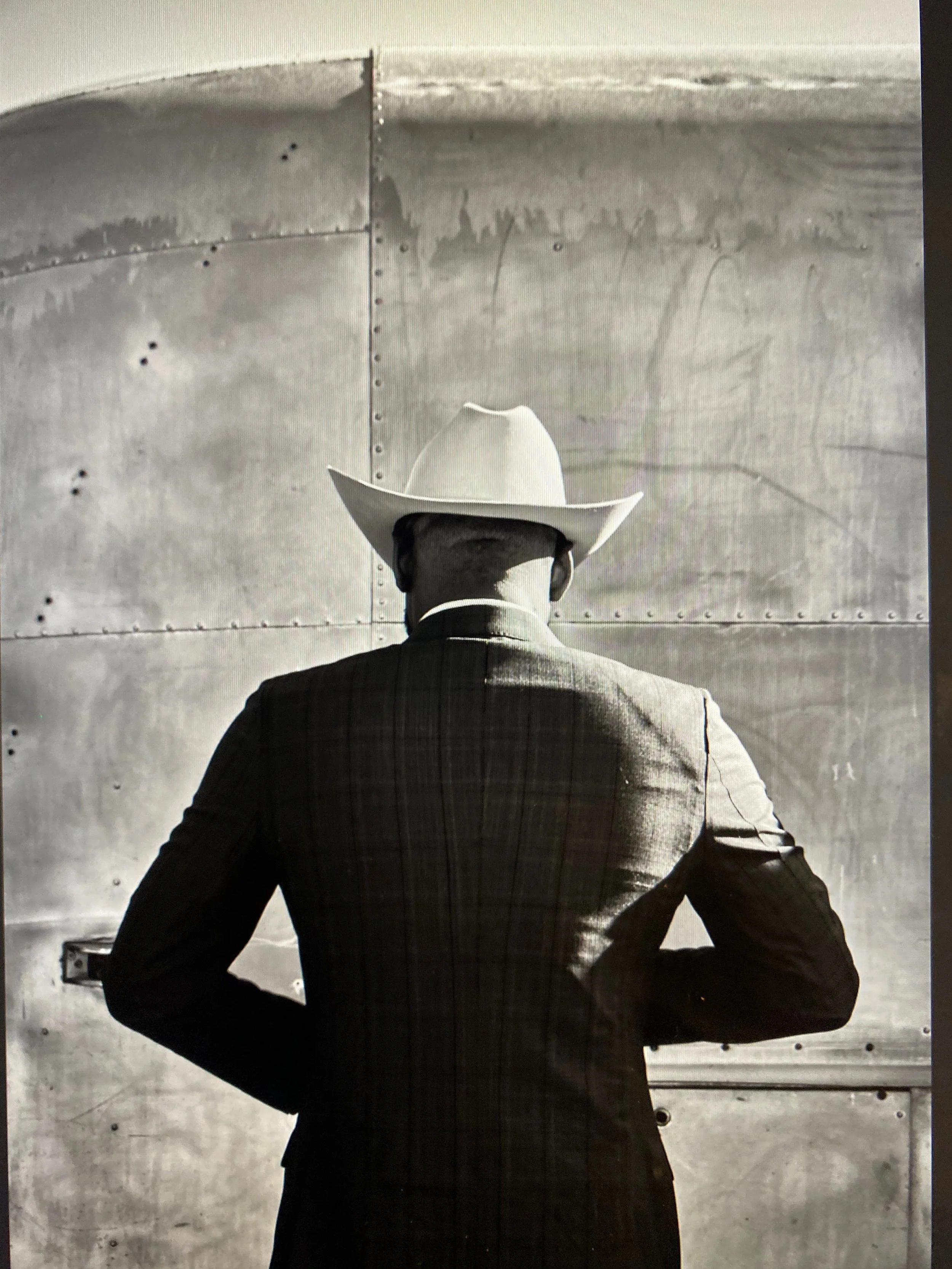 A person in a suit and wide-brimmed cowboy hat standing with their back to the camera, looking at a metal wall of an Airstream.