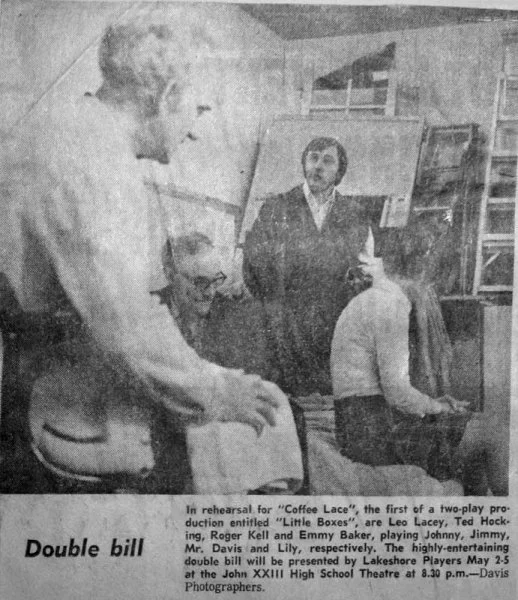 Black and white photo of four people in a drama rehearsal, with one woman standing and three others sitting, including a woman with glasses and a man with a beard, in front of a stage with paintings and props.