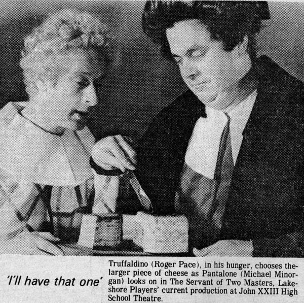 Two actors on stage, a woman on the left with curly hair and a man on the right with dark, thick hair, are engaged in a scene involving a large piece of cheese. The woman is smiling, and the man appears focused. They are dressed in period costumes. T