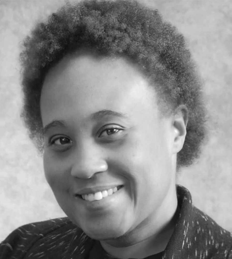 A black and white portrait of a smiling woman with short, curly hair, wearing a patterned jacket.