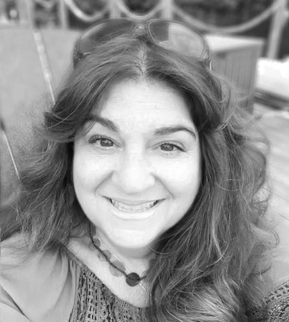 A woman with long wavy hair smiling, wearing sunglasses on her head and a necklace, with outdoor furniture and a railing in the background.