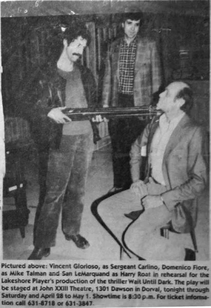 Three men in a room with one seated and two standing. The seated man is in a suit, while the standing men are dressed casually. One standing man holds a gun pointed at the seated man's head. The background includes a wooden cabinet and some indistinc