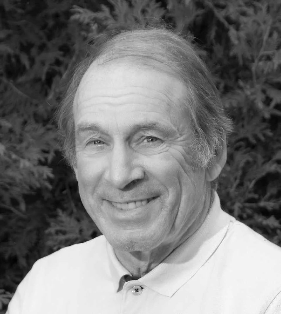 A smiling man with light hair and wearing a collared shirt, outdoors with blurred foliage in the background.
