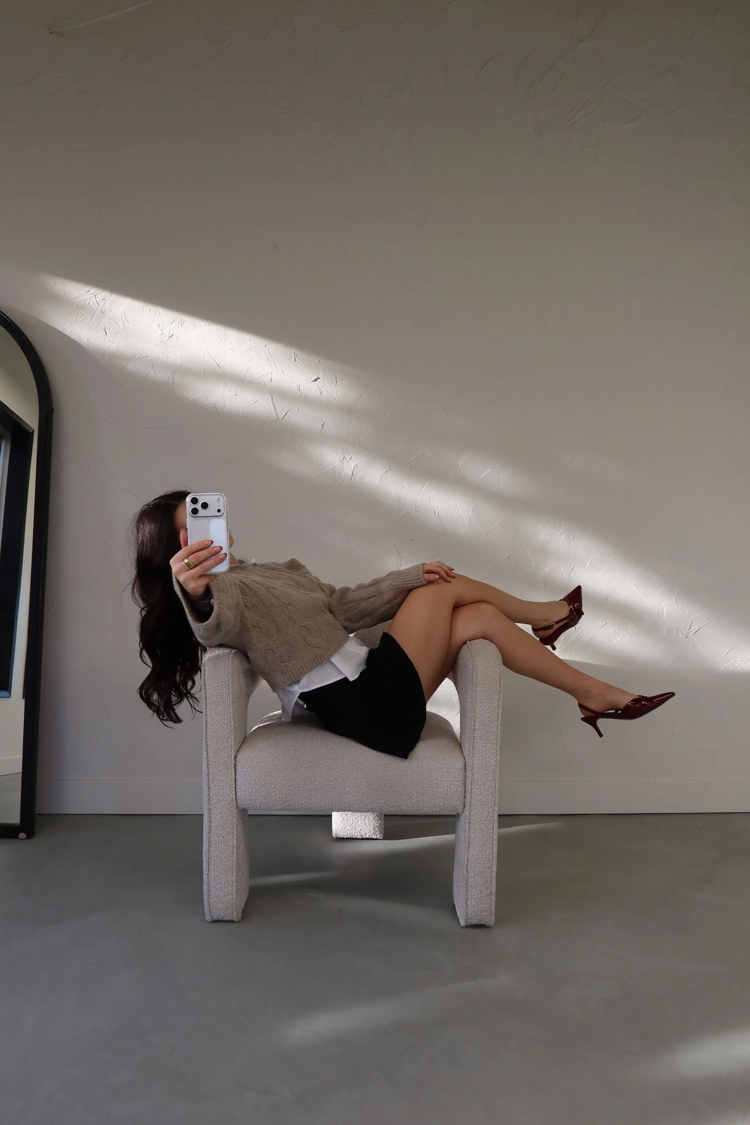 A woman is sitting on a beige armchair, taking a mirror selfie. She is wearing a beige knitted sweater, a white shirt, a black skirt, and burgundy high heels. Her face is obscured by the phone.