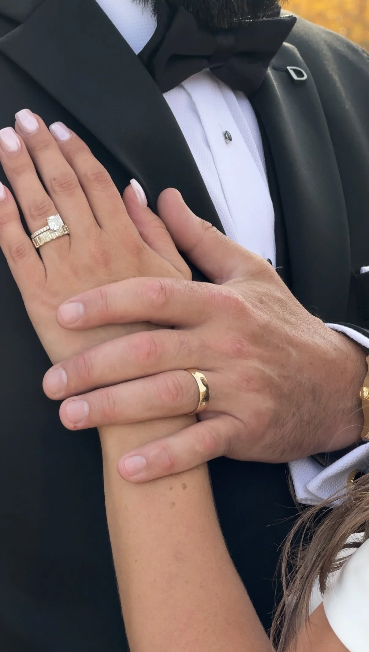 A man and woman wearing wedding rings, with the man's hand on top of the woman's hand, both dressed in formal attire including a tuxedo and rings.