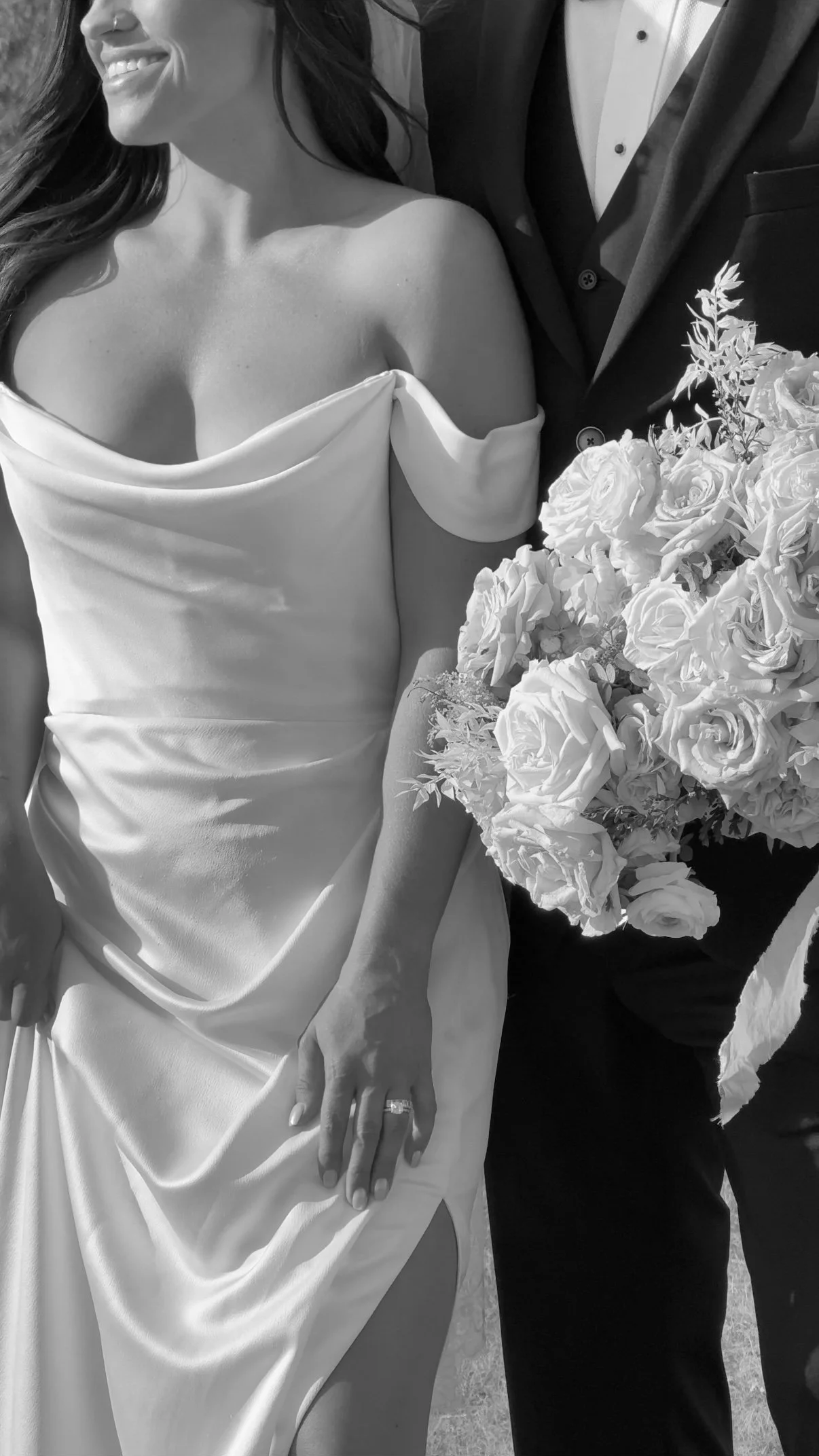 A woman in a wedding dress is smiling, showing her ring finger with a wedding band, holding a bouquet of flowers next to a man in a suit.
