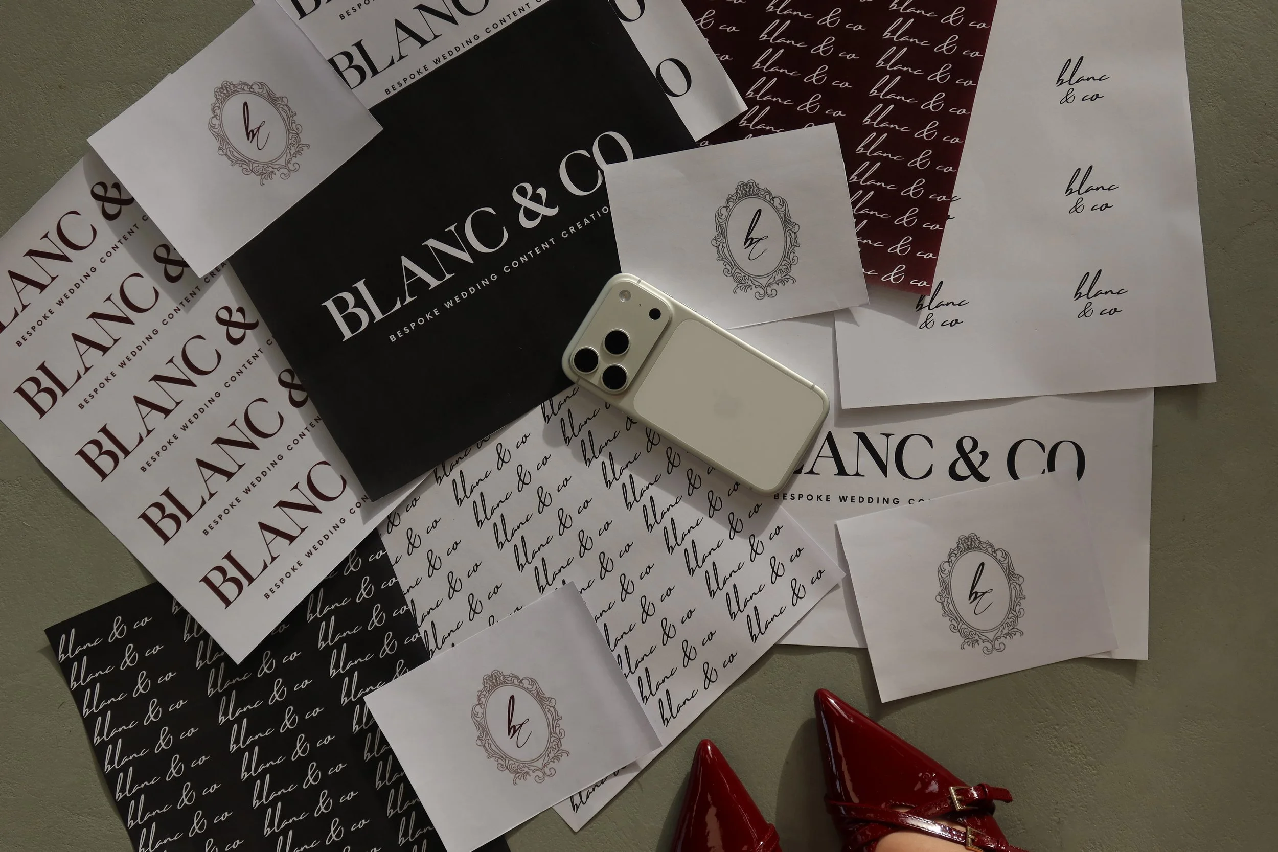 Various sheets of paper with branding and script designs for 'blanc & co,' along with a white iPhone, a red pair of shoes, and a beige surface.