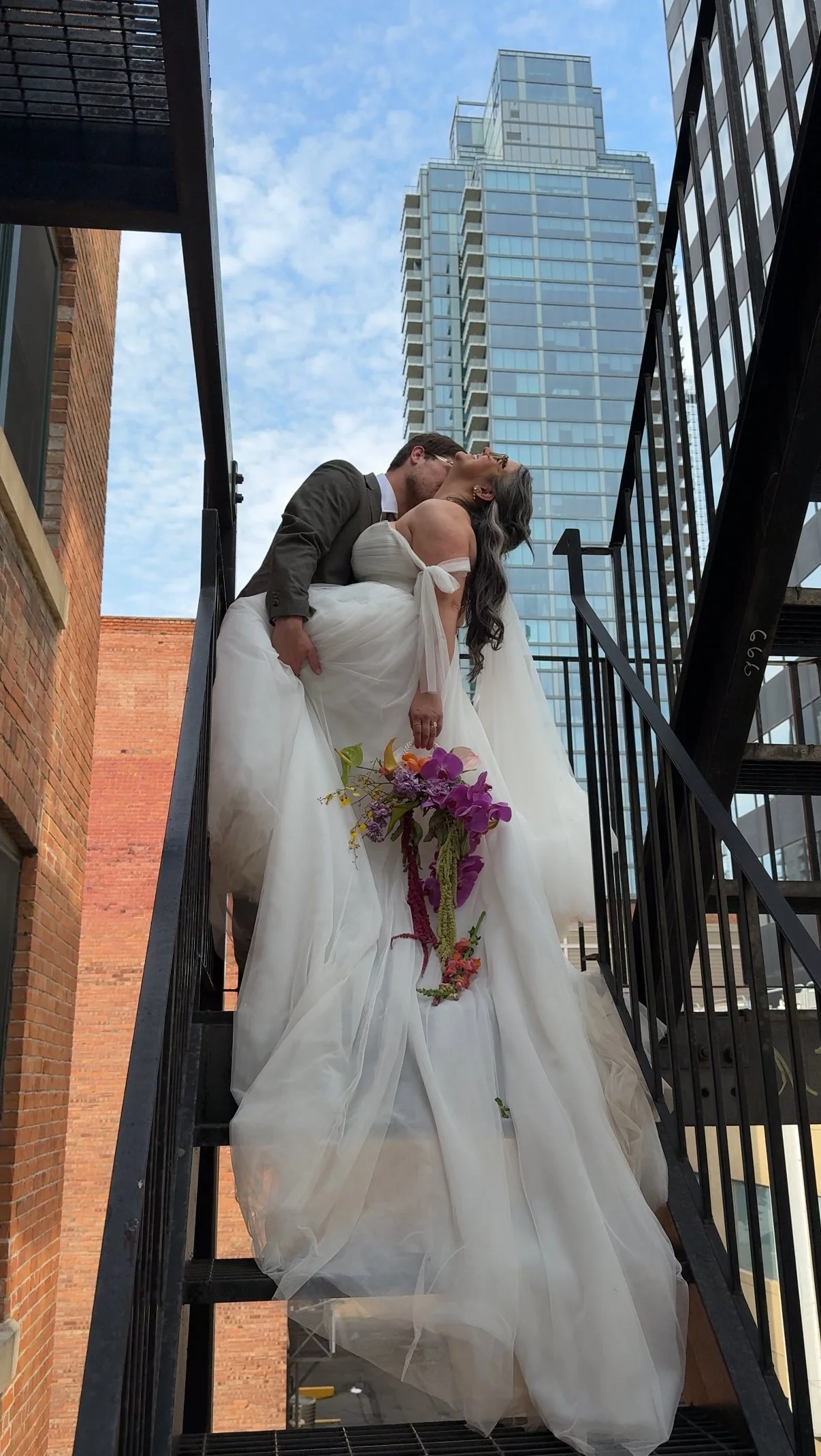 A bride and groom share a kiss on a fire escape; the bride is holding a bouquet of purple and pink flowers, and tall modern buildings are visible in the background.