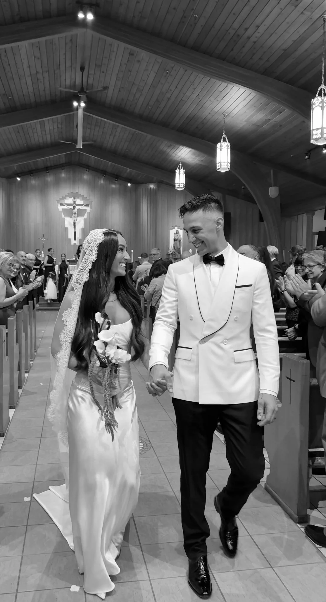 Black and white photo of a newly married couple walking down the aisle of a church, holding hands and smiling at each other. The bride wears a satin wedding dress and veil, holding a small bouquet, while the groom is dressed in a tuxedo with a bow tie. Guests are clapping and smiling in the background.