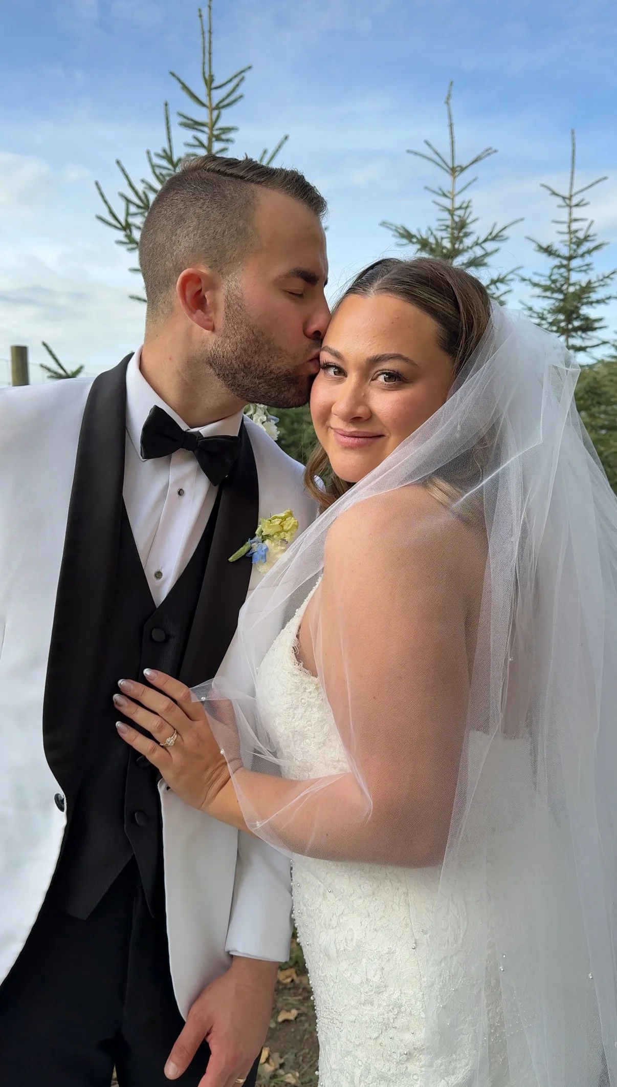 A bride and groom on their wedding day outdoors. The groom is kissing the bride on the forehead. The bride is smiling, wearing a lace wedding dress and veil. The groom is dressed in a tuxedo with a black bow tie.