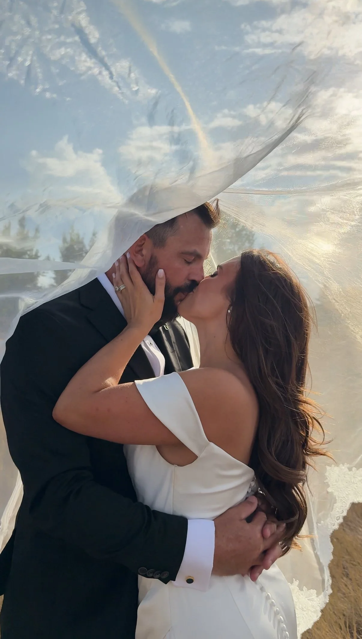 A bride and groom share a kiss outdoors with a veil overhead reflecting the sky and trees.