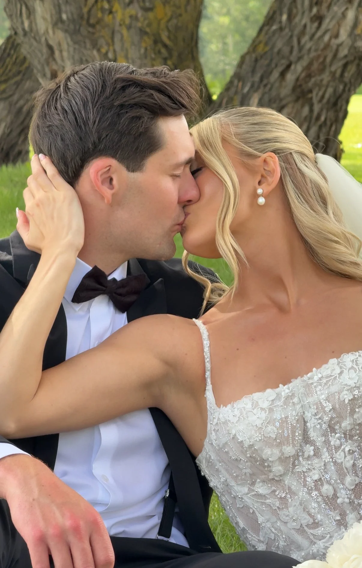 A bride and groom kiss outdoors during their wedding ceremony, with trees in the background.