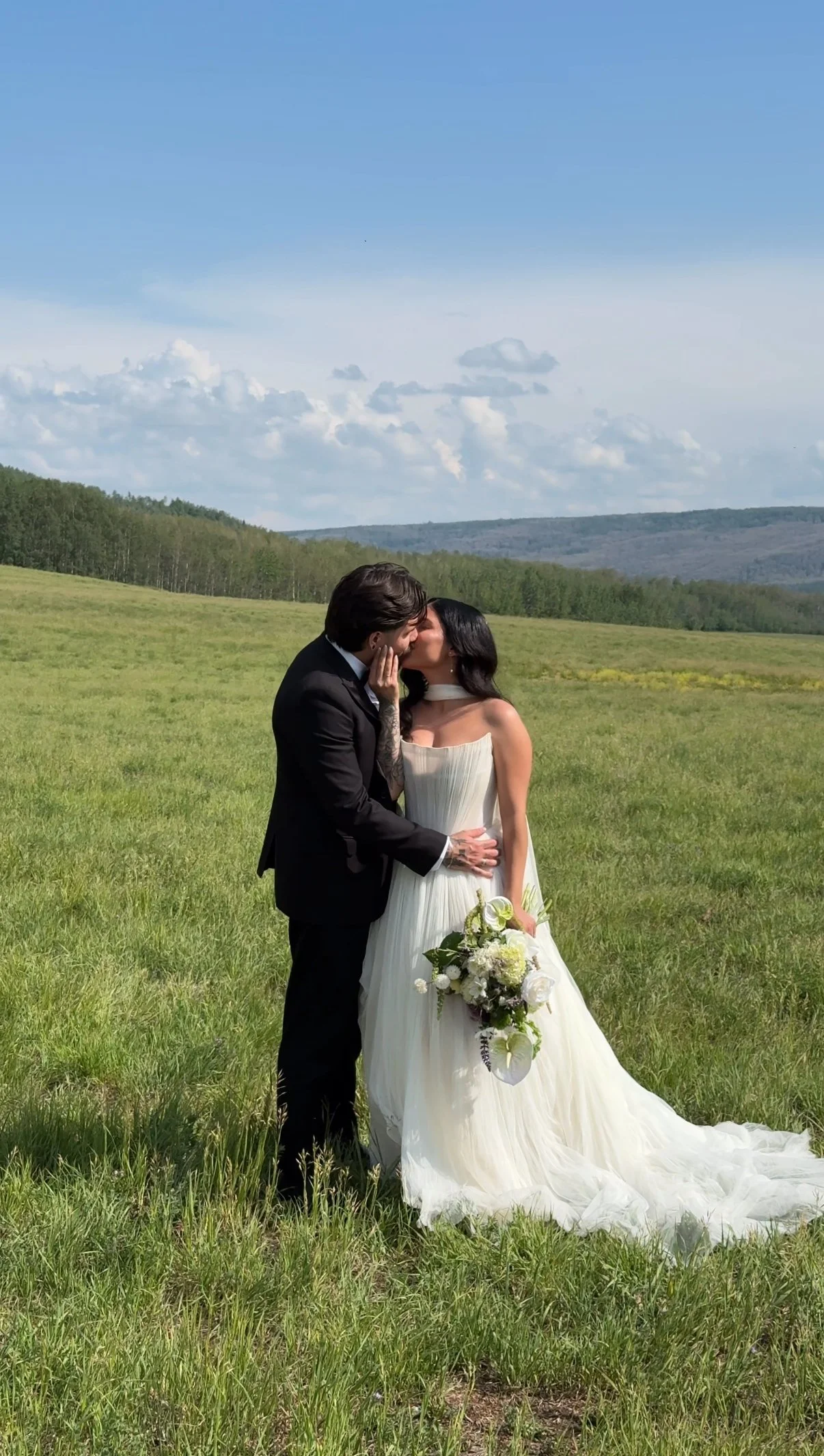 A bride and groom share a kiss in a grassy field with rolling hills and a blue sky with clouds in the background.