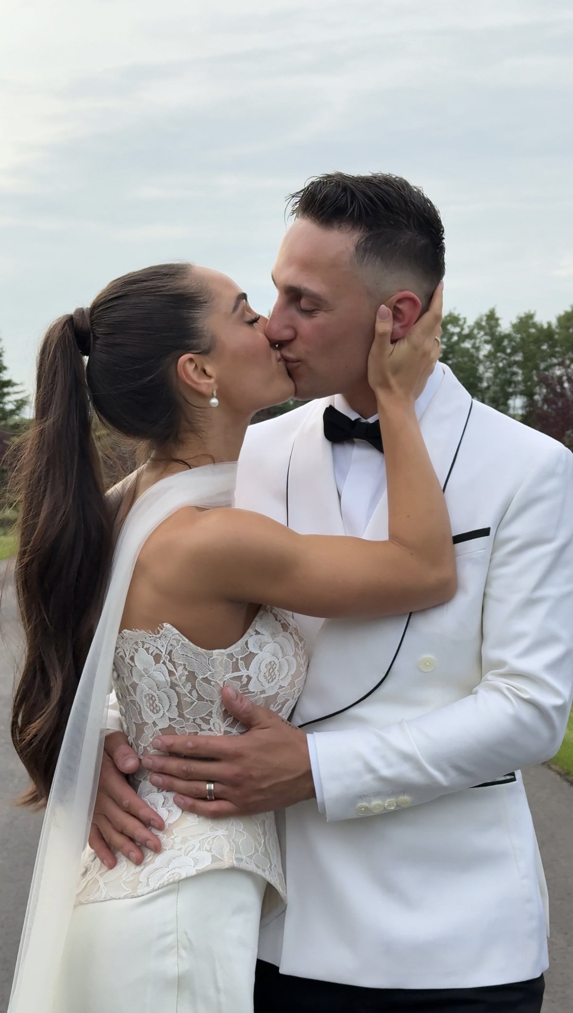 A bride and groom kiss outdoors, with the bride holding the groom's face and the groom embracing her waist, on a cloudy day.