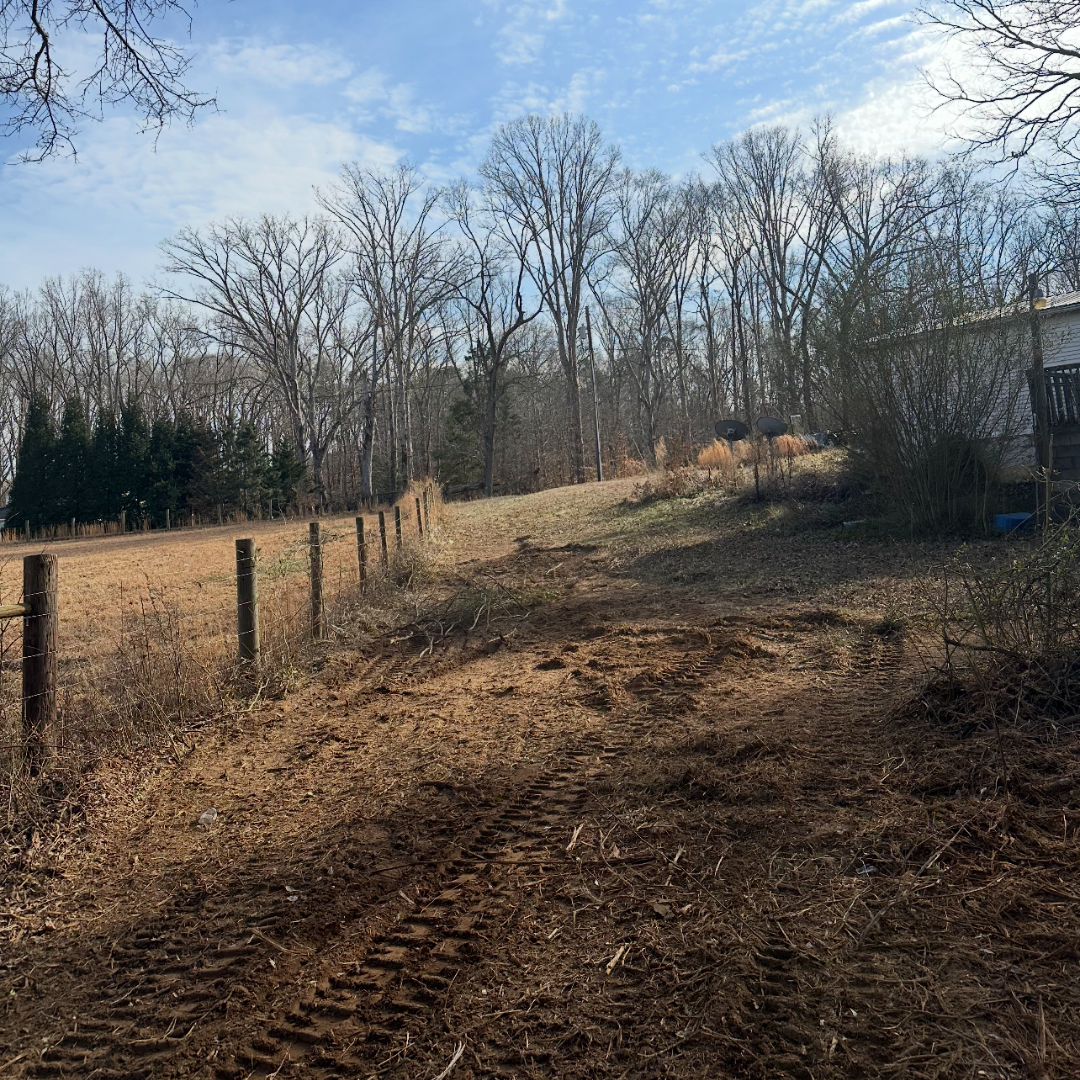 A dirt pathway with tire tracks in a rural backyard, bordered by a wooden fence on the left and a white house with satellite dishes on the right, with leafless trees and a partly cloudy sky in the background.