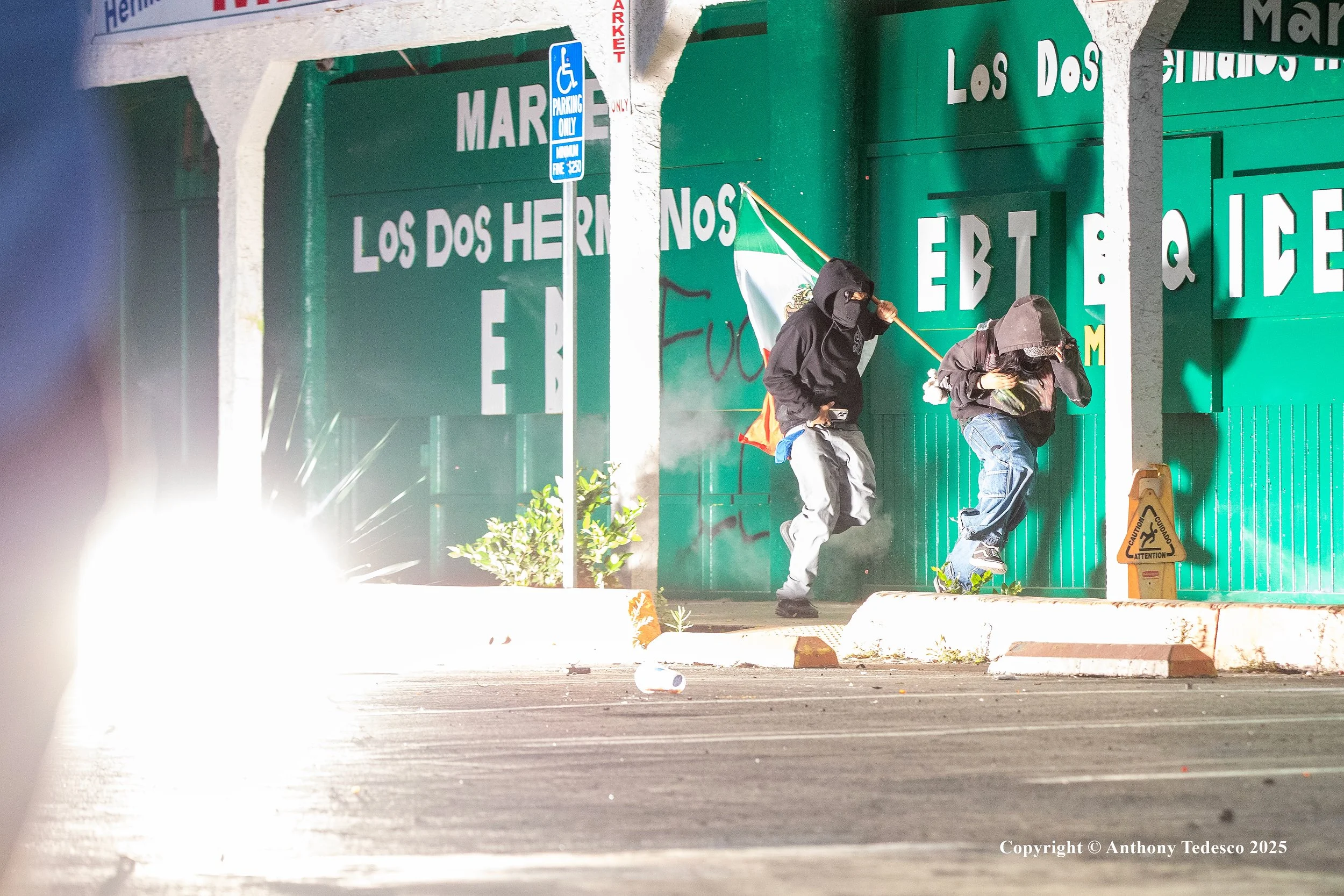 Two individuals dressed in black hoodies and jeans, with one holding a Mexican flag, running away from tear gas at night in front of a green wall with 'LOS DOS HERMANOS' written on it, near a handicap parking sign.