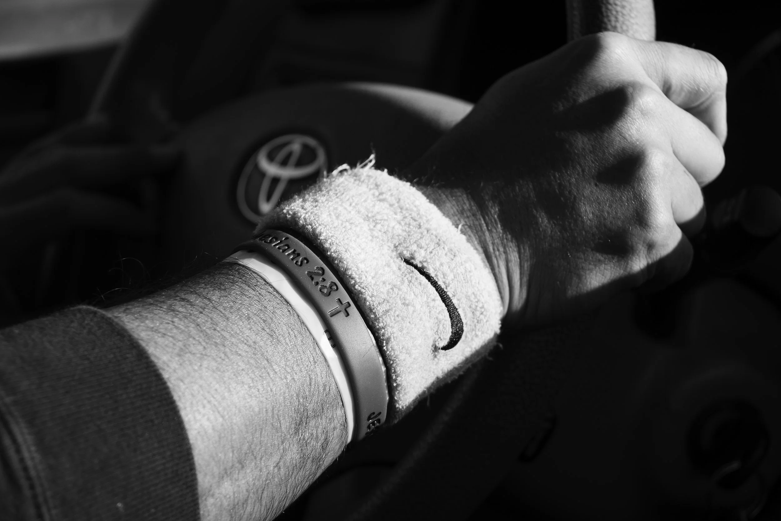 A person's arm with a fuzzy wristband and a bracelet, holding a steering wheel inside a car, with the Toyota logo visible on the steering wheel.