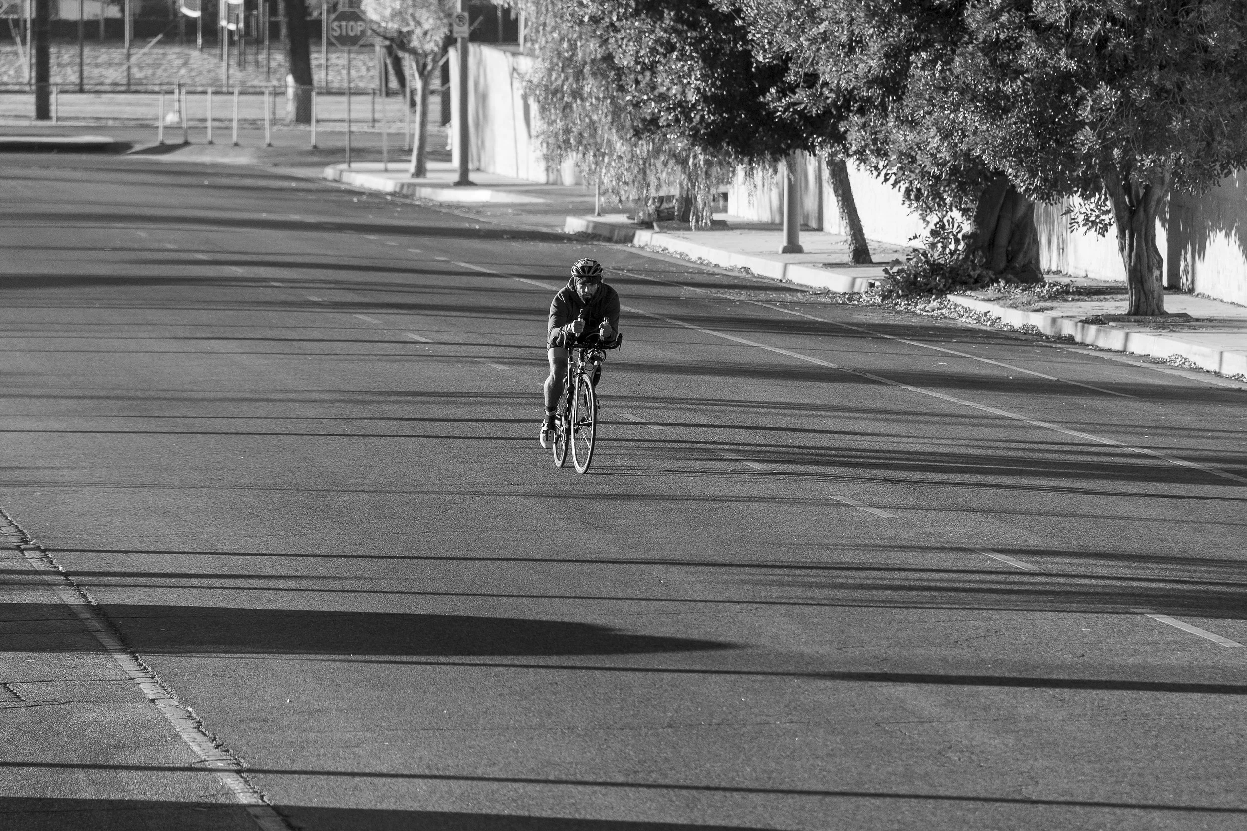 A person riding a bicycle on an empty street with trees and sidewalk in the background, in black and white.