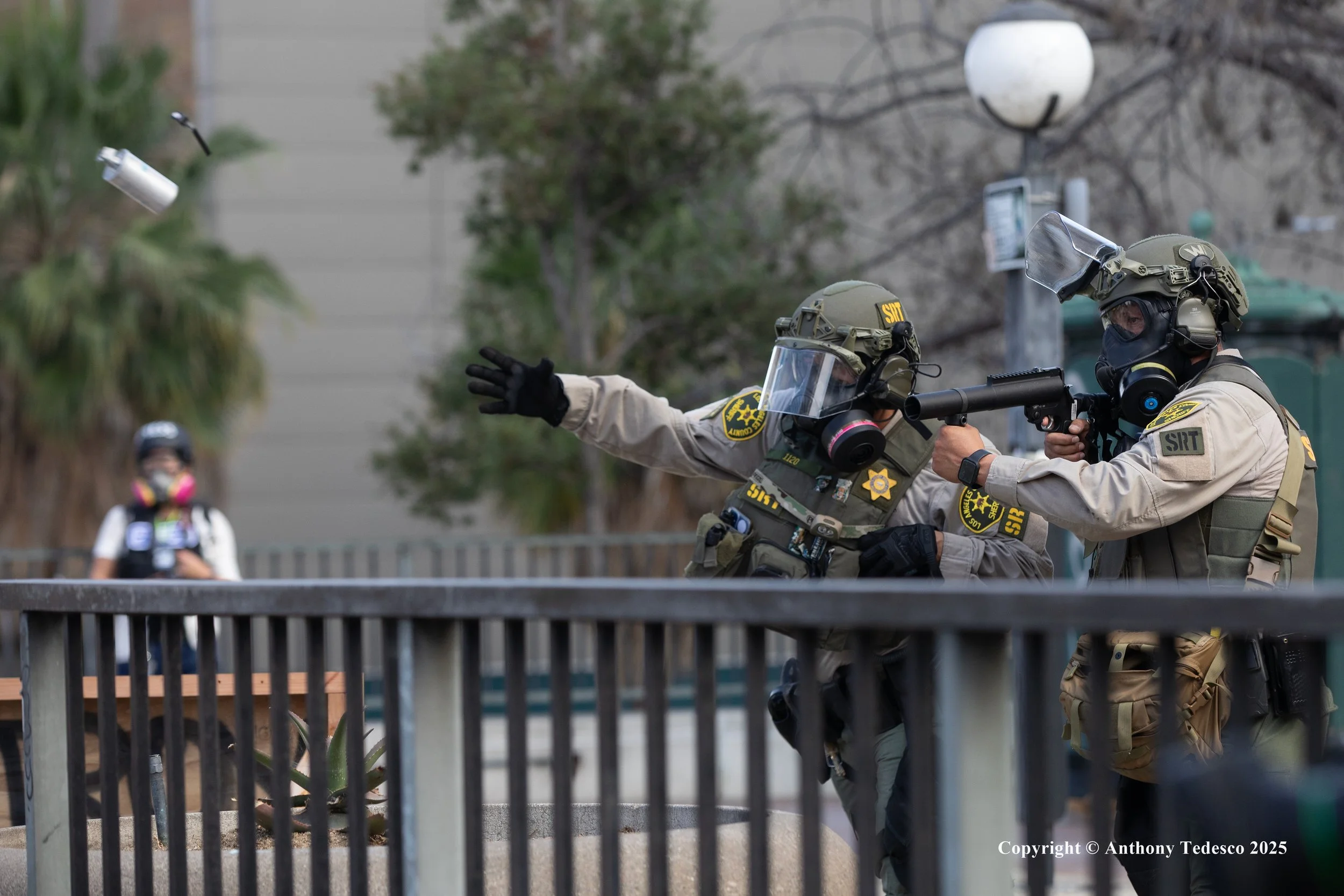 Two law enforcement officers in tactical gear aiming a gun at a flying object, while a woman in a mask observes in the background.