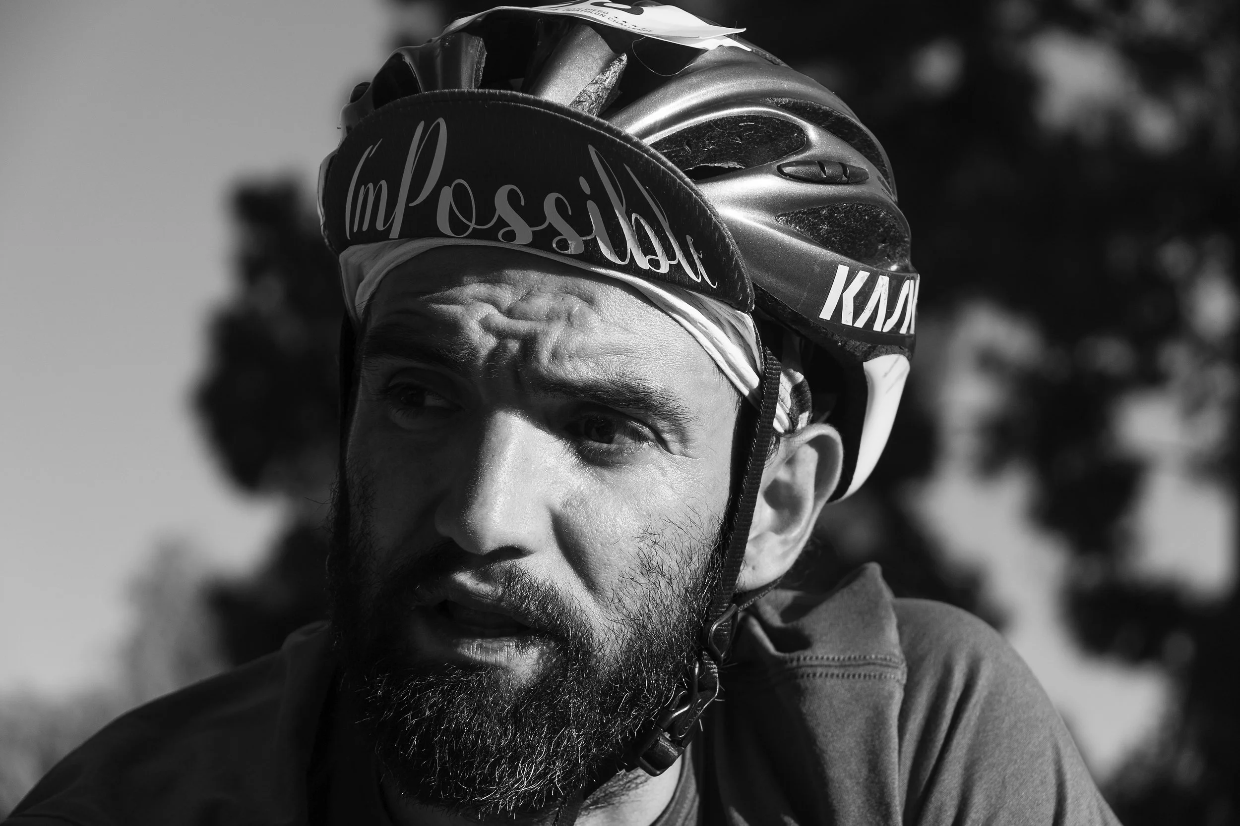 A man wearing a cycling helmet and headband, with the brand 'KASK' and the words 'I'm Possible' on his gear, with a serious expression and outdoor background.
