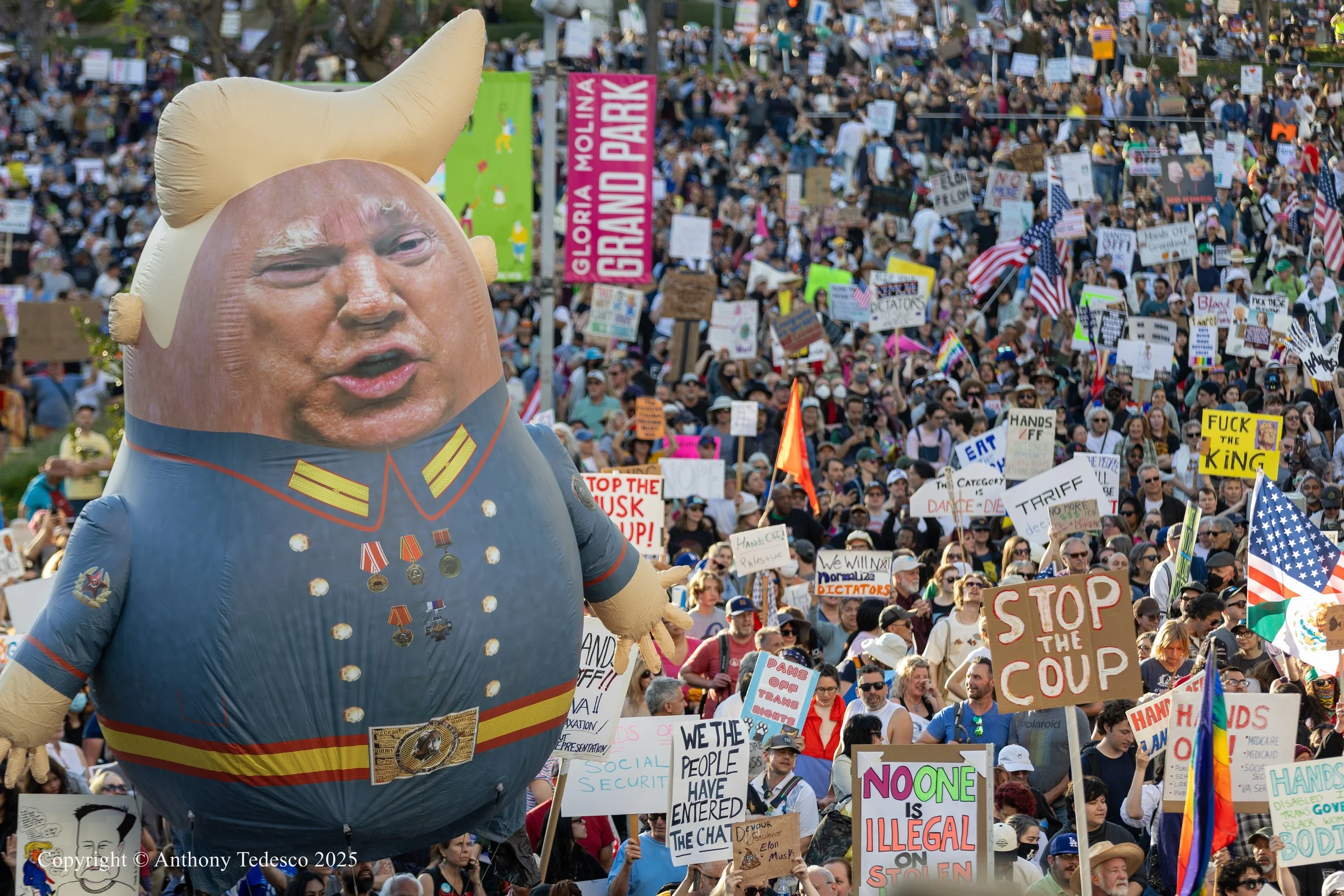 Large inflatable balloon resembling a caricature of Donald Trump in a military uniform amidst a crowded protest with many smaller signs and flags.