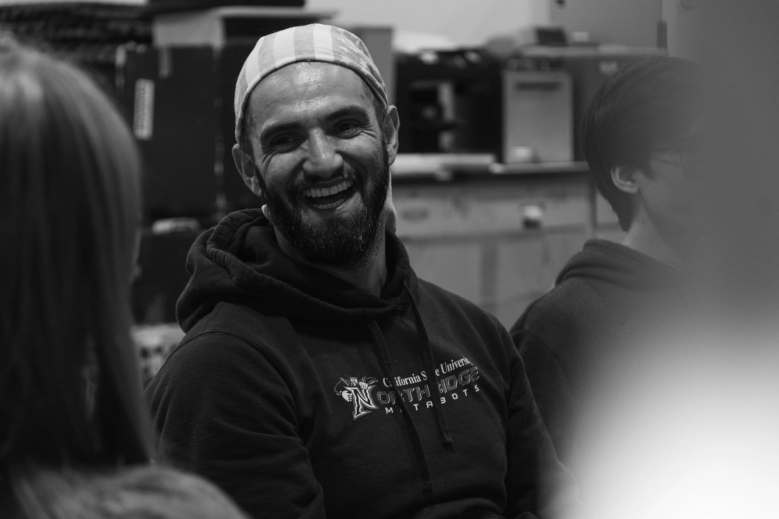 A man with a beard wearing a hoodie and a headband, smiling and talking to two other people in an indoor setting.