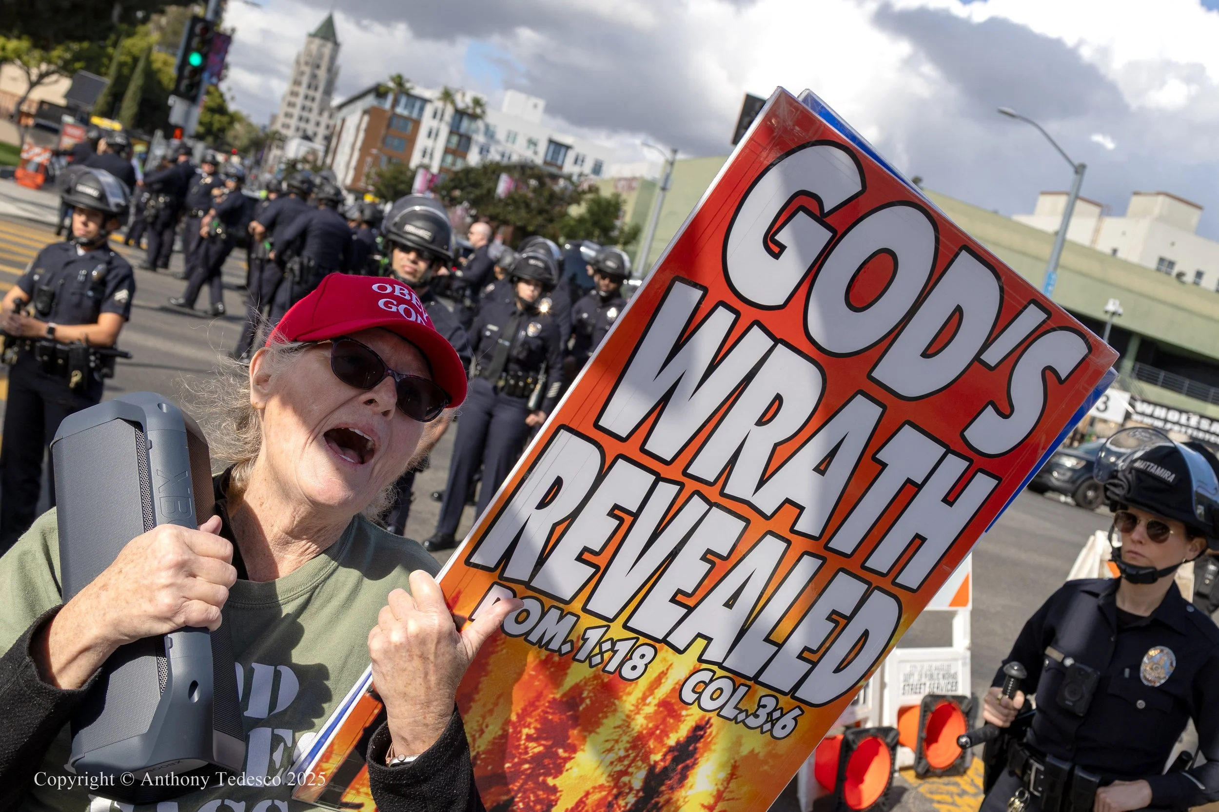 Elderly woman protesting with a sign that reads 'God's Wraith Reached' at a demonstration against COVID restrictions, with police officers in riot gear in the background.
