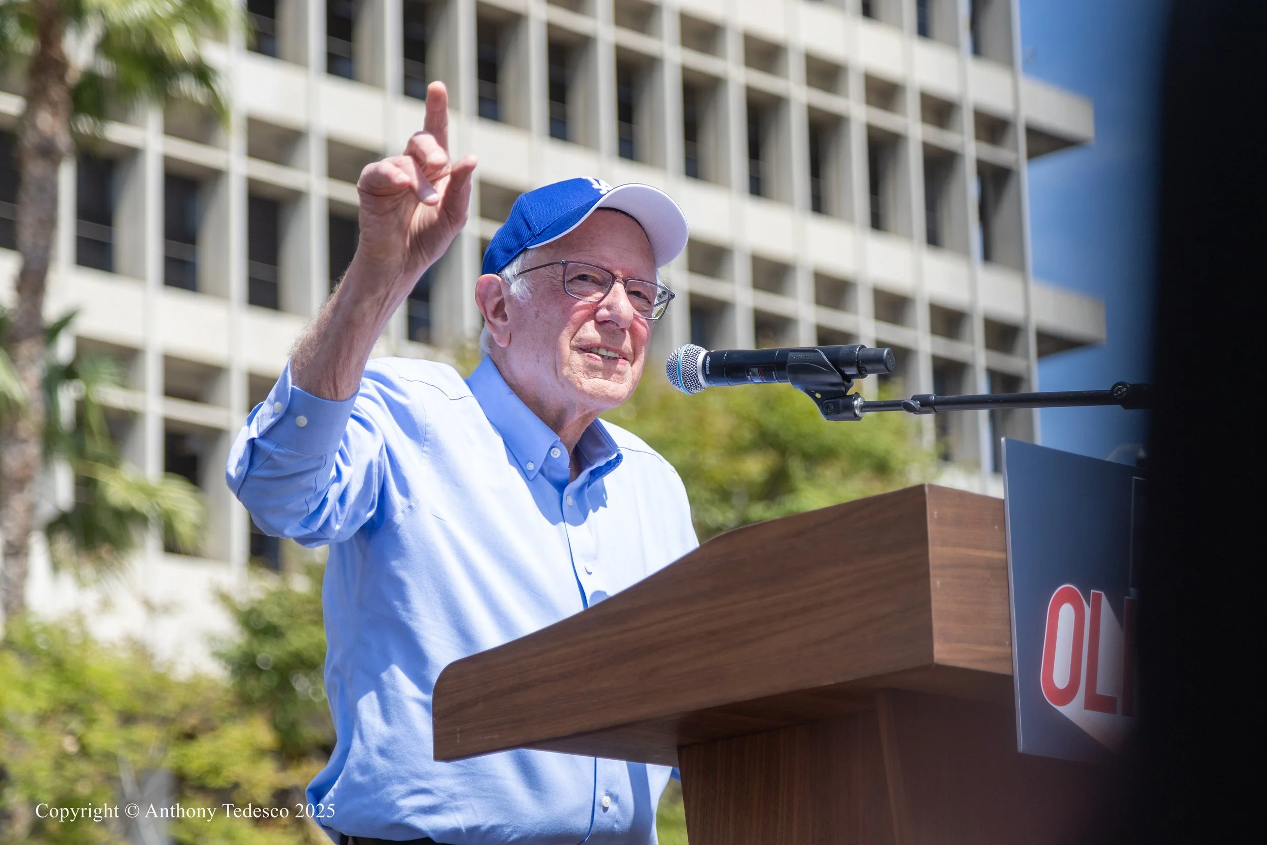 Bernie Sanders is speaking at a podium with a microphone in front of him. He is gesturing with his right hand, raising his index finger. Behind him, there are trees and a modern building with a grid-like facade.
