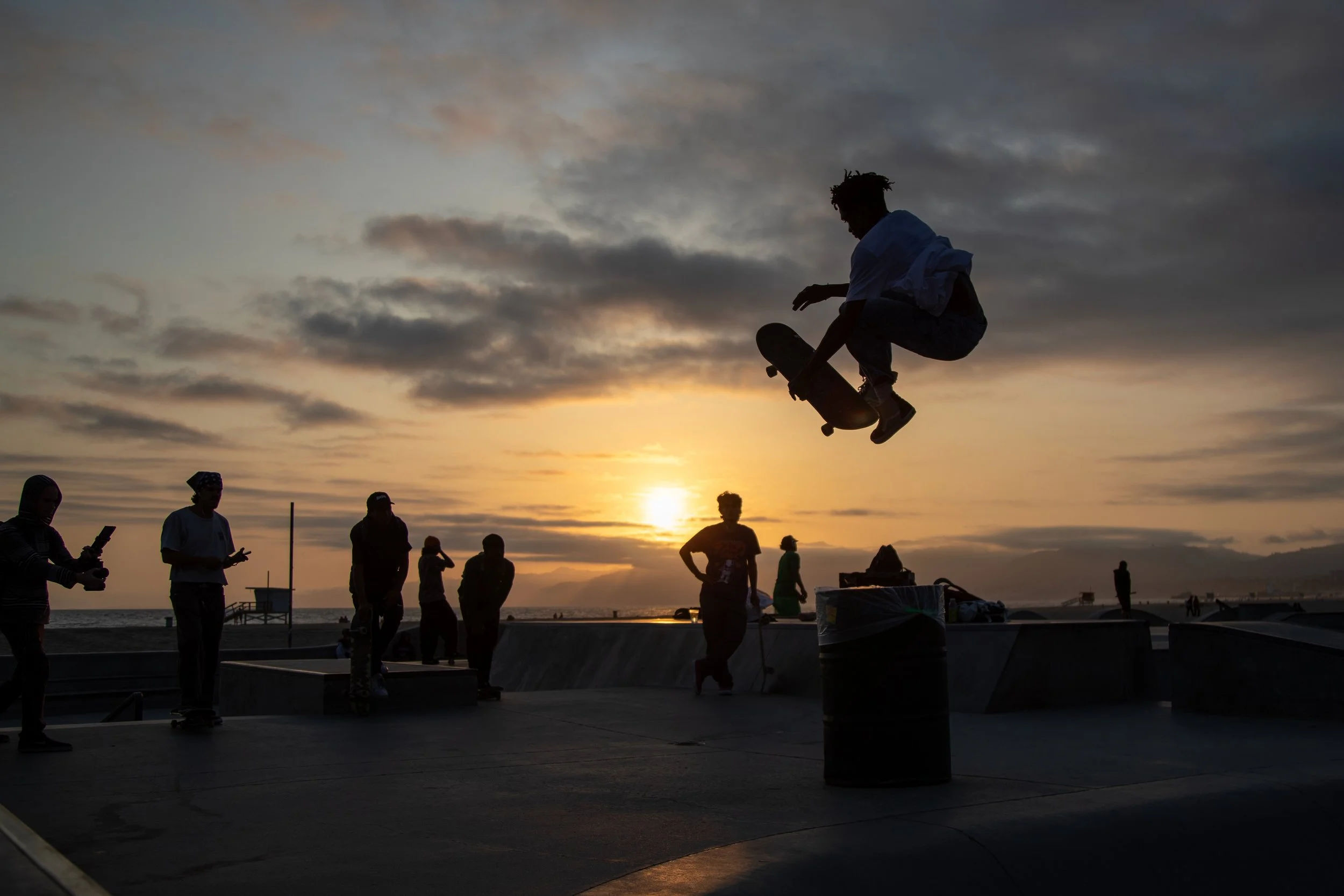 Skateboarder performing a jump at sunset at the skatepark with people in the background.