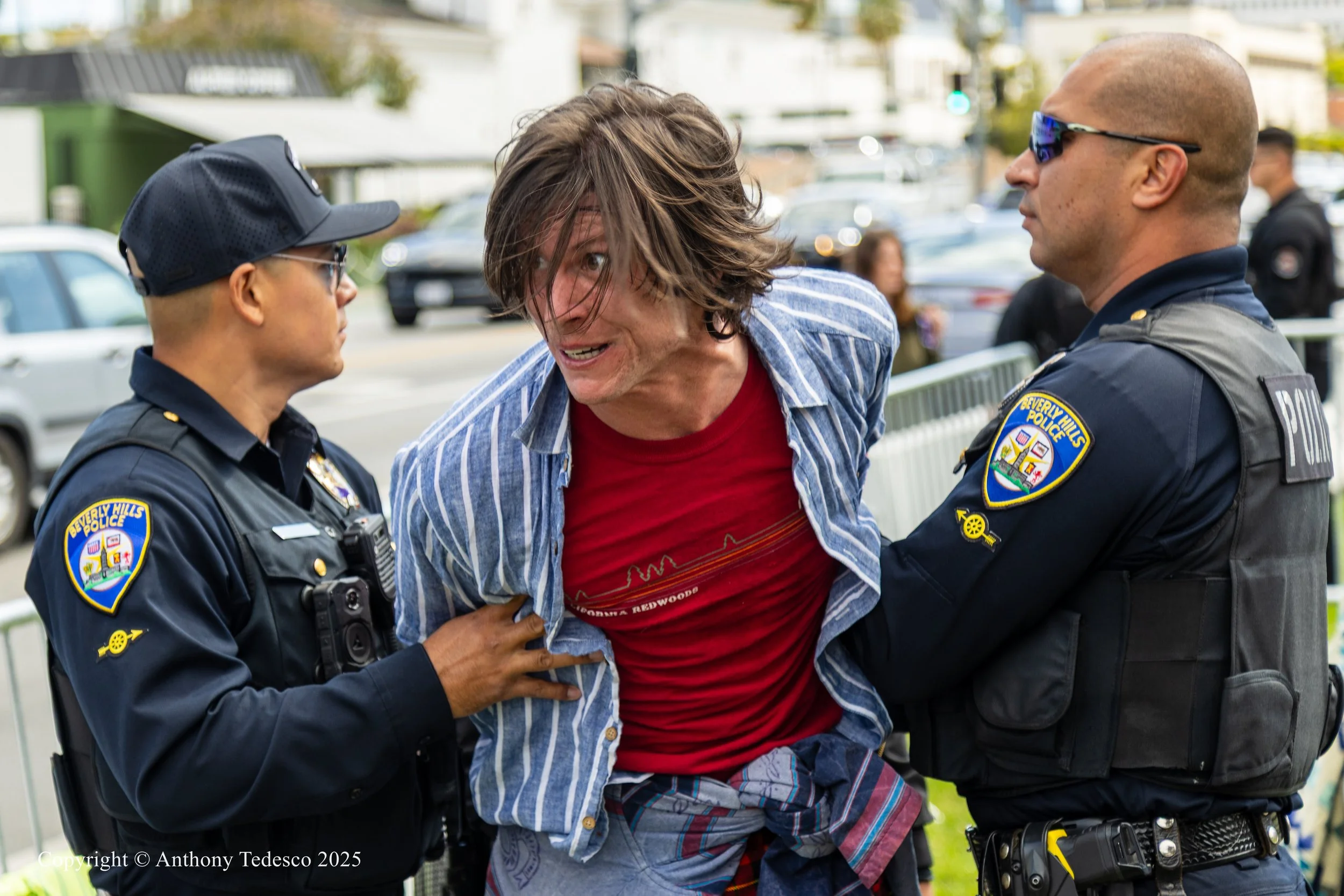 A distressed man in a red T-shirt and striped shirt being restrained by two police officers with badges and dark uniforms during a protest or demonstration on a city street.