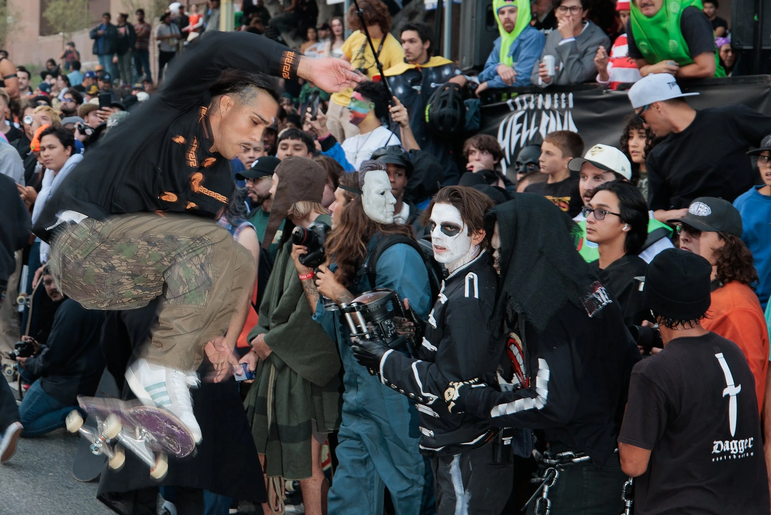 A crowd of people gathers at a protest or rally. Several individuals are wearing face paint and masks, and some are dressed in costumes or casual clothing. One person is climbing onto another, and others are holding cameras and cameras. In the backgr