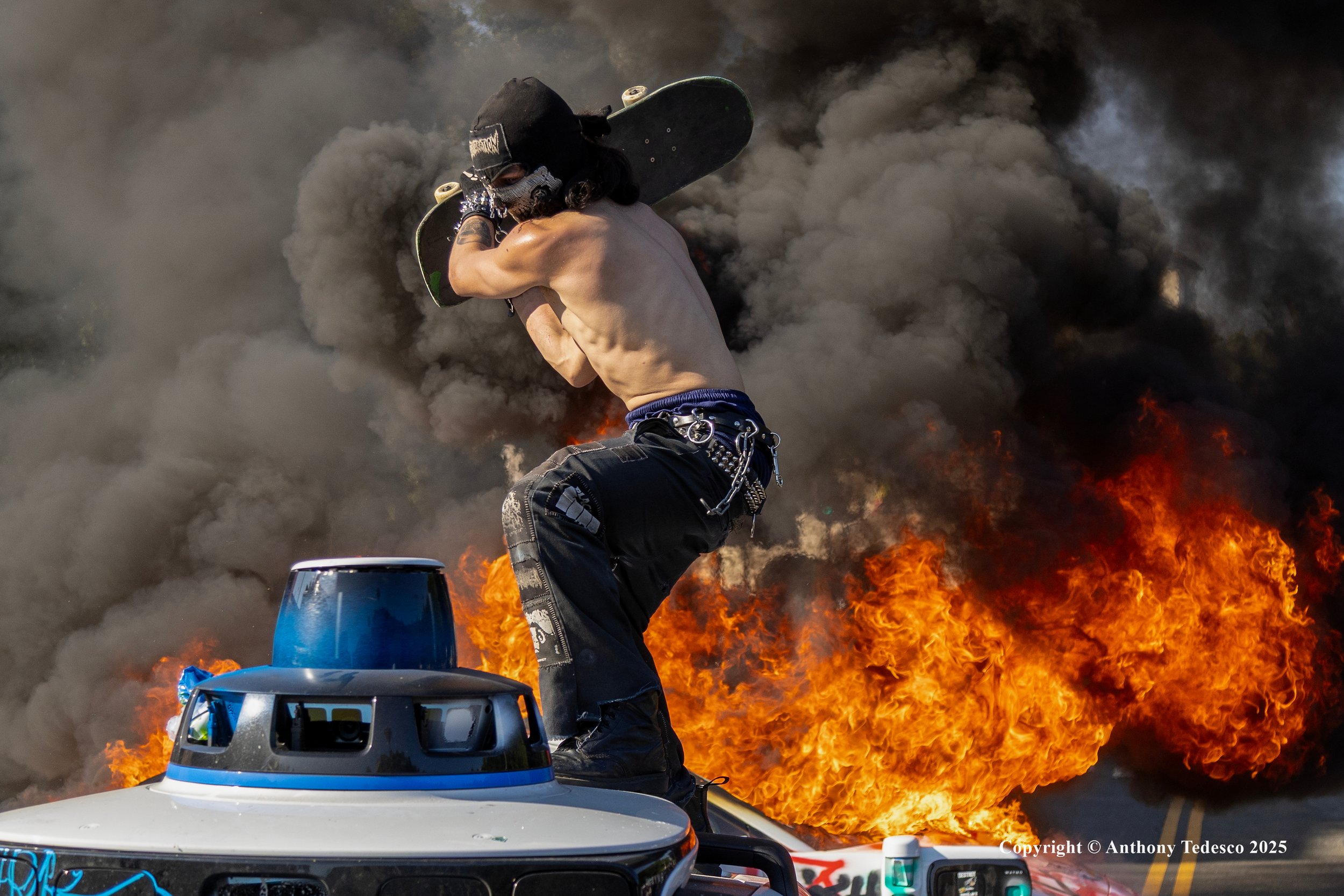 A shirtless man wearing a mask and black pants, holding a skateboard, is standing on a vehicle amid a large fire with thick black smoke in the background.