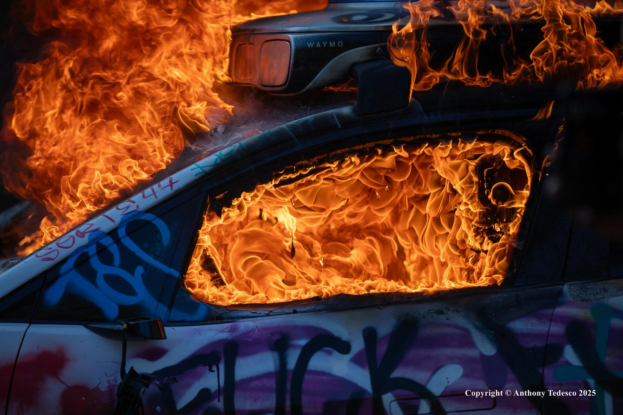 A car on fire with flames engulfing the entire vehicle, which is covered with graffiti and spray paint.