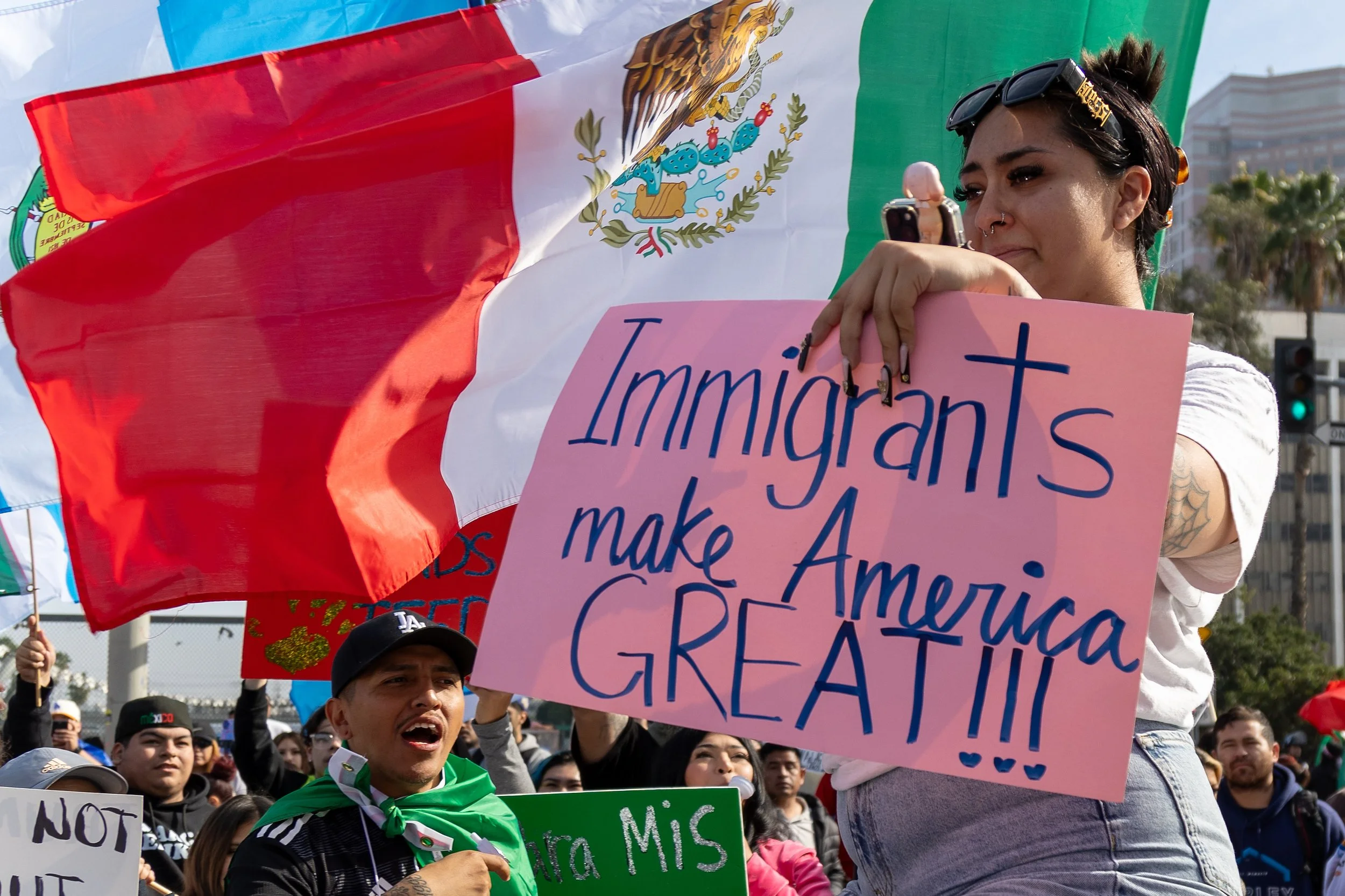 A woman holding a pink sign that says 'Immigrants make America GREAT!!!' is at a protest or rally. There are flags, including the Mexican flag, and a crowd of people around her.