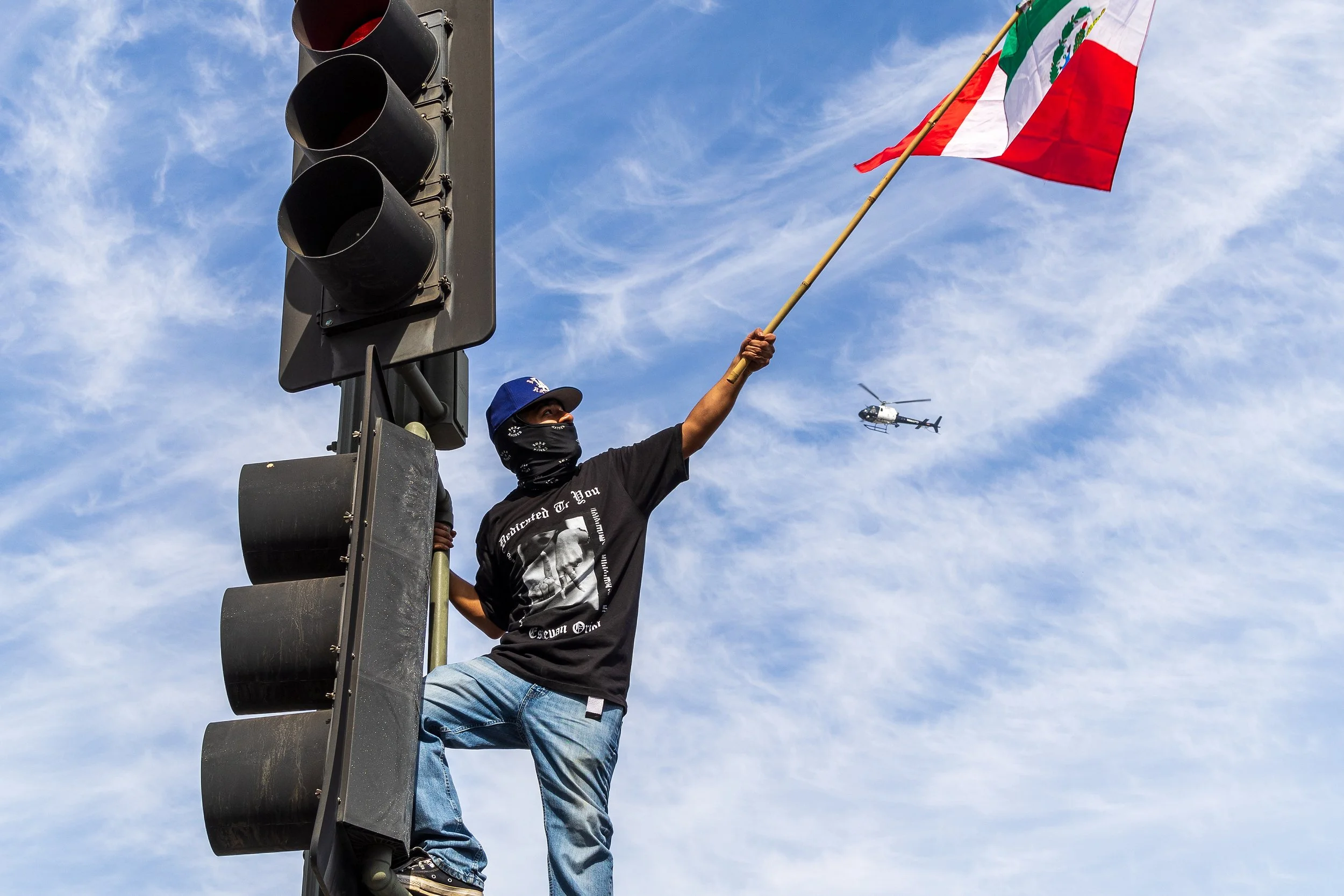 Person standing on a pole holding a Mexican flag during a protest, with a helicopter flying in the background.
