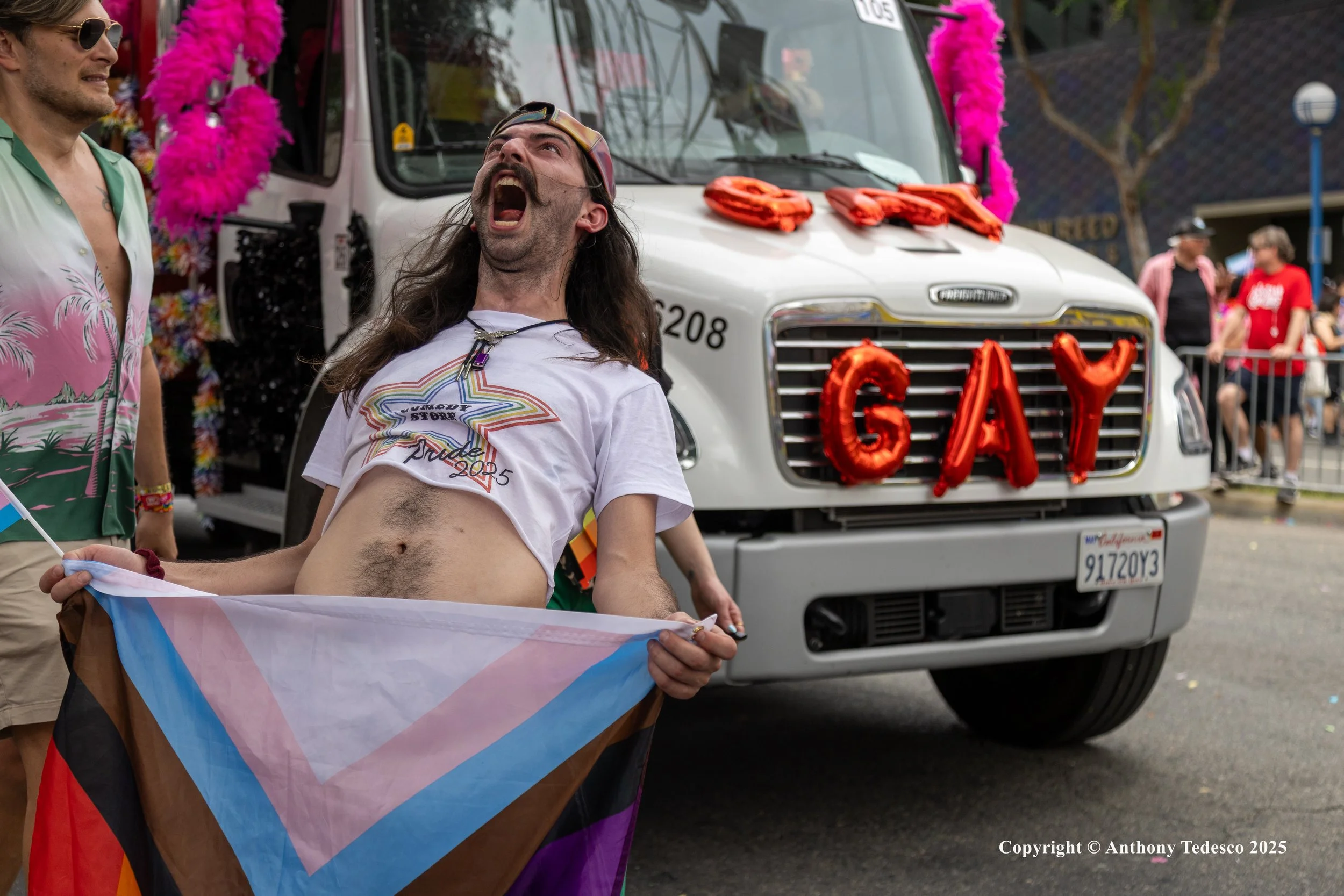 A person at a pride parade holding a rainbow flag and wearing a T-shirt with a star design. They are dancing in front of a decorated vehicle with the word 'GAY' spelled out in red balloons. The vehicle is a truck with pink decorations and a license p