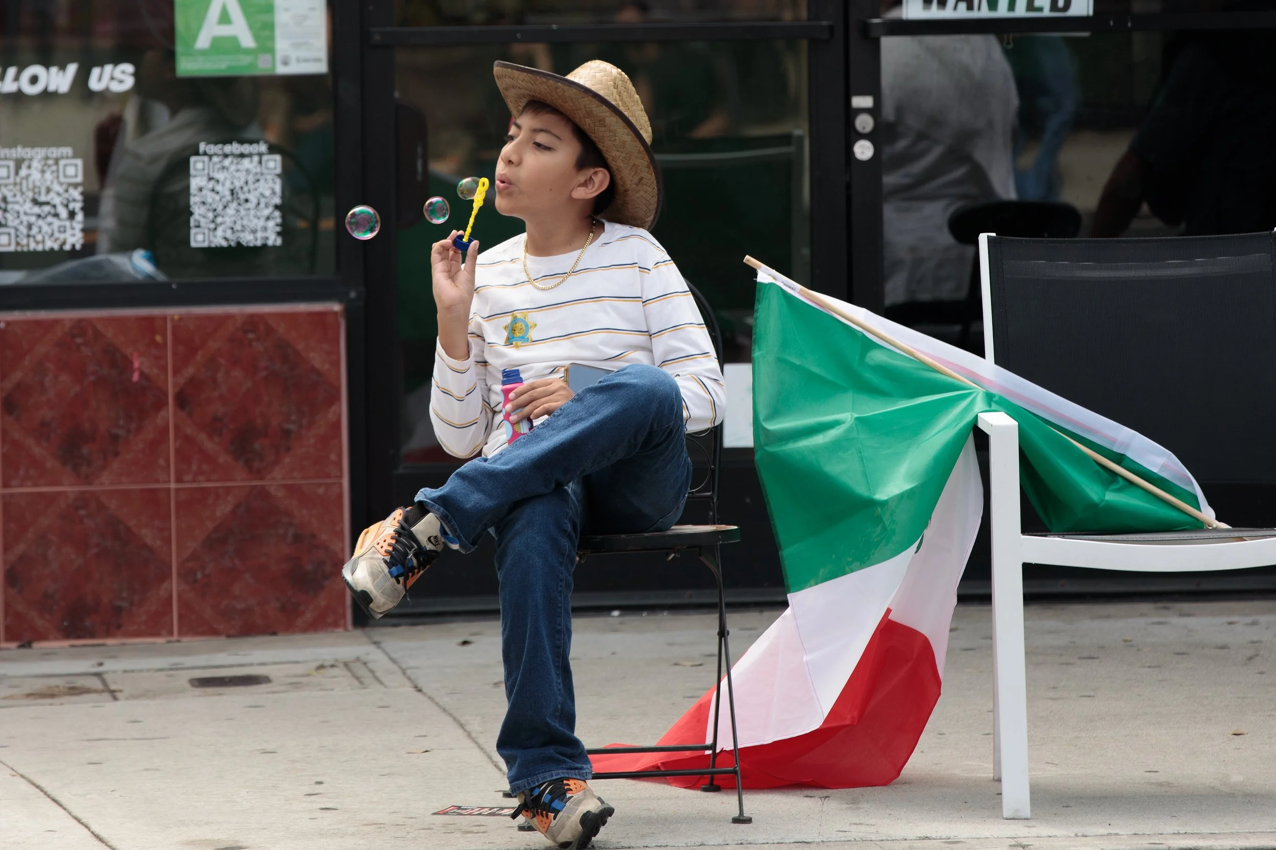 A young boy wearing a straw hat, striped shirt, and jeans sitting on a black chair outside a building, blowing bubbles, with an Italian flag placed over the chair.