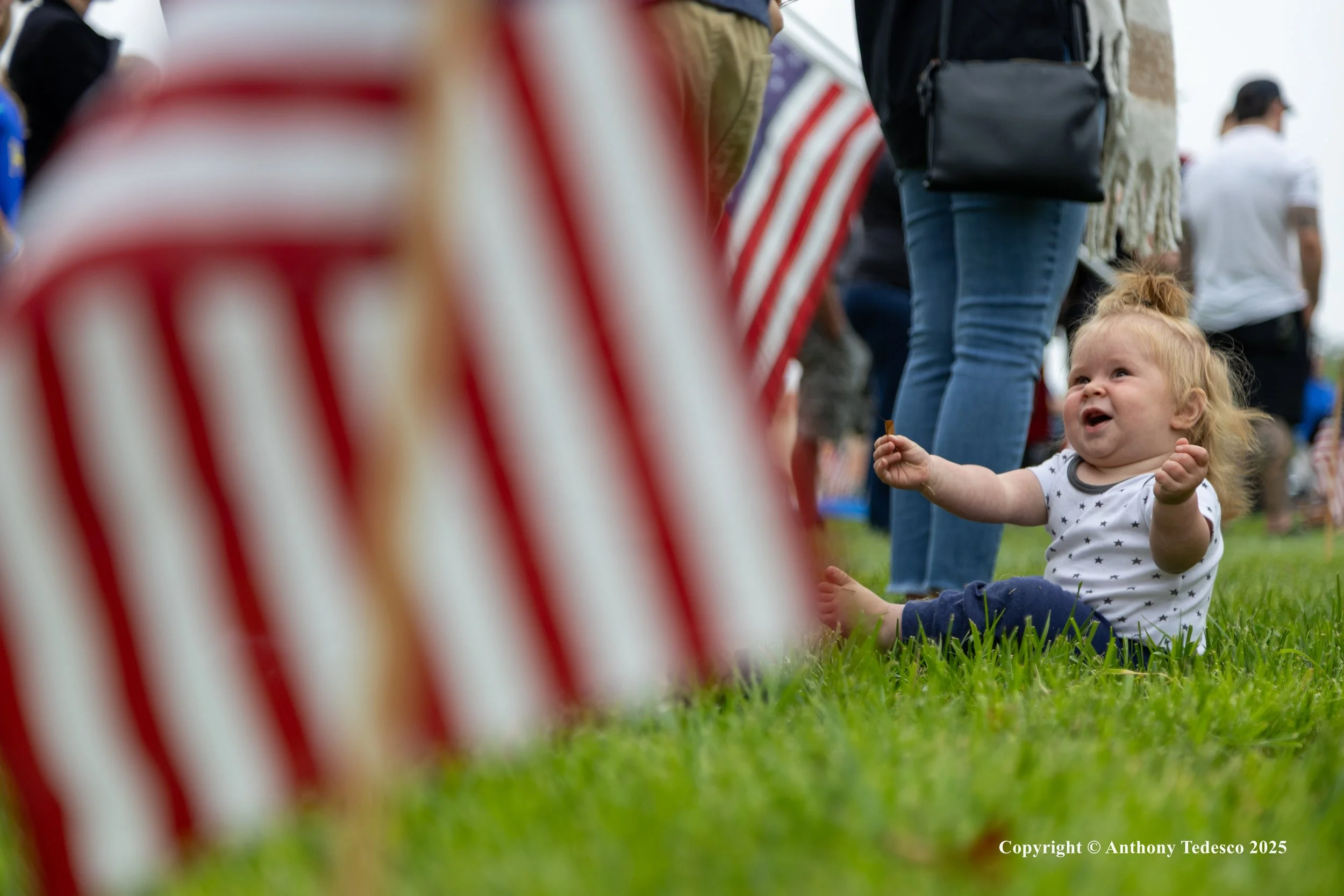 A happy baby sitting on the grass at an outdoor event, with American flags and people in the background.