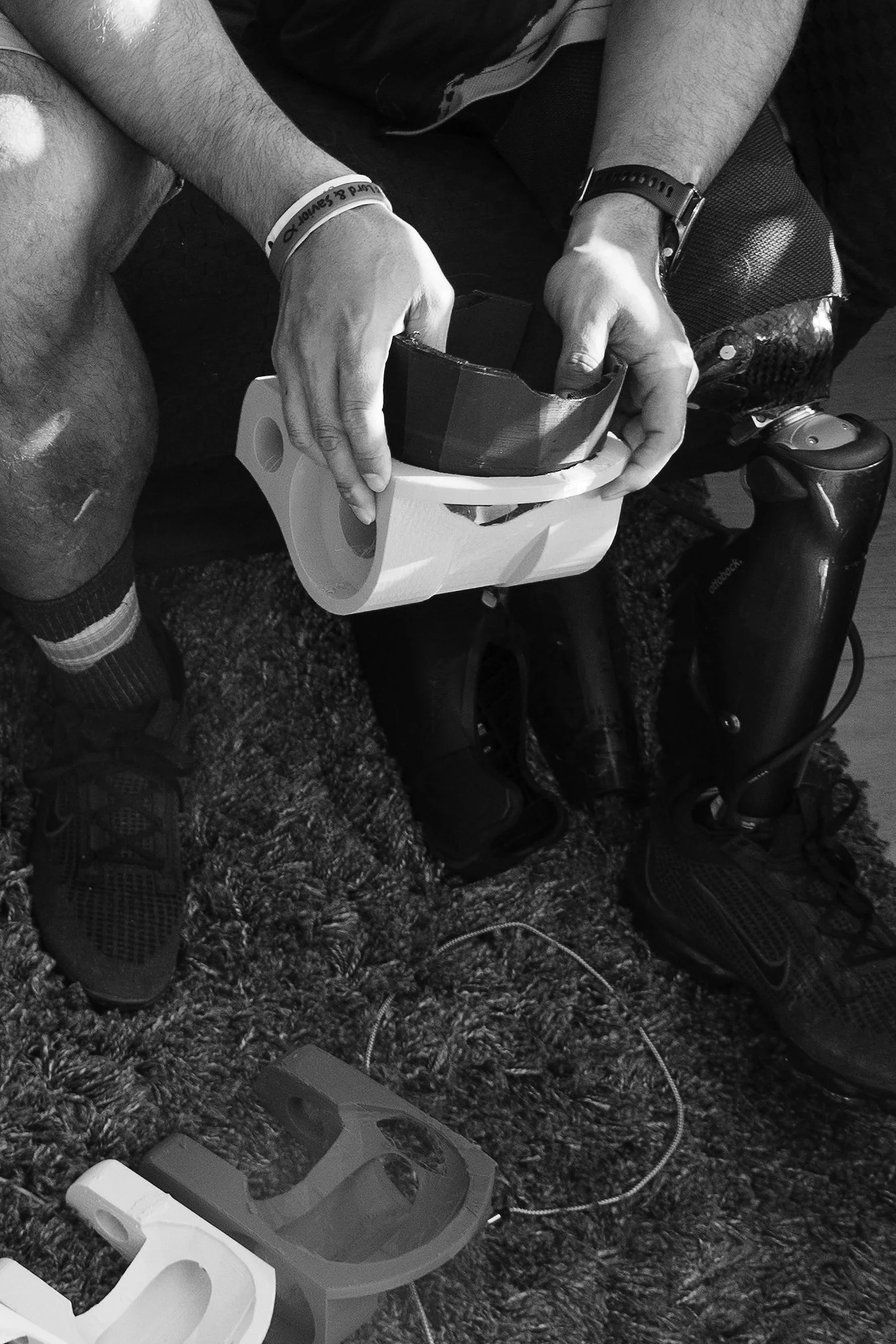 Person seating on a textured carpet, wrapping a large roll of black tape with a utility knife, surrounded by shoes, a backpack, and headphones.