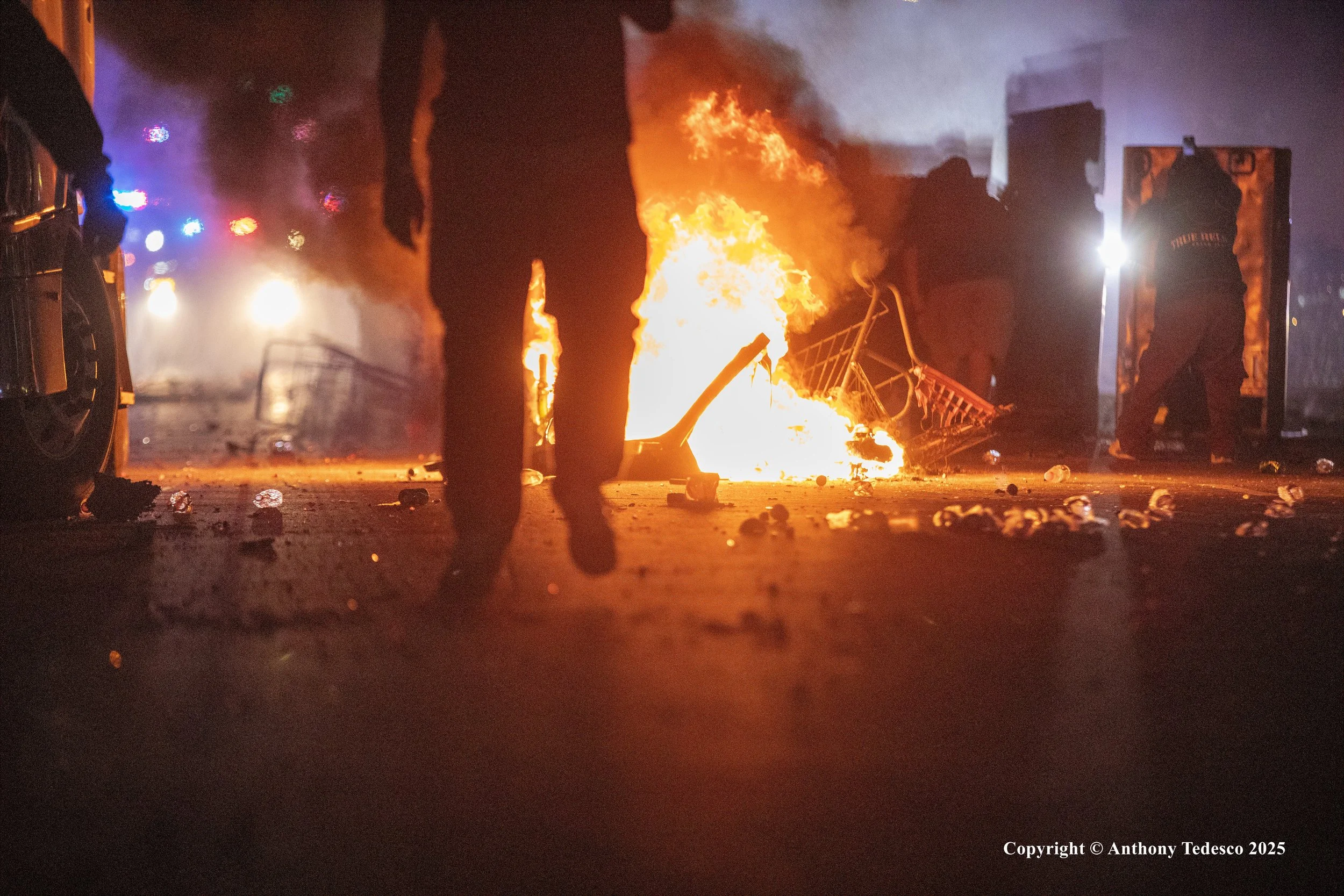 A car is on fire on a city street at night, with flames and smoke. Debris is scattered on the ground, and two people are visible near the fire, one standing and one walking away. Emergency lights are visible in the background.