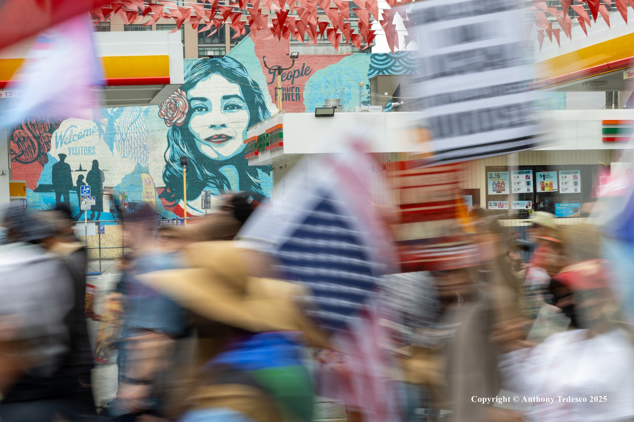 Blurred scene of people walking past a 7-Eleven store, with a large mural of a woman's face and the words "People Power" in the background.
